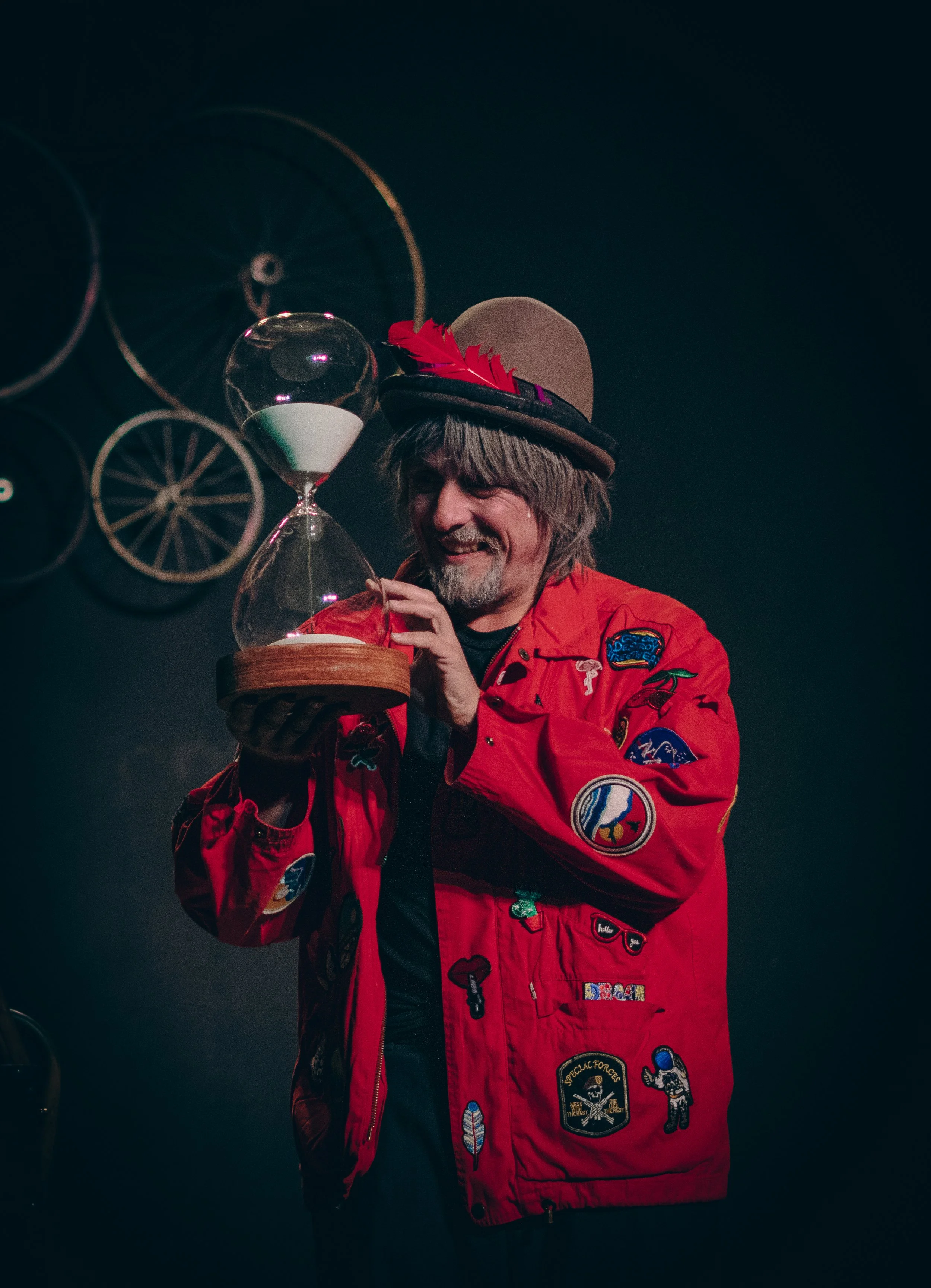 Hombre con chaqueta roja decorada con parches, sombrero con pluma roja, sosteniendo un reloj de arena en un escenario oscuro.