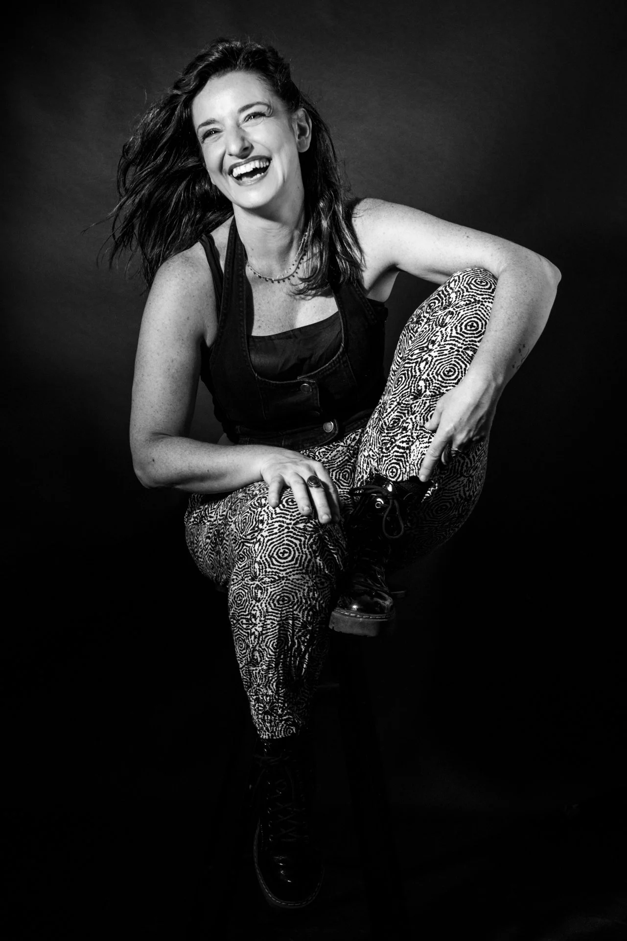 Mujer sonriendo, cabello suelto, vestida con ropa oscura y pantalones con patrón geométrico, en un fondo negro.