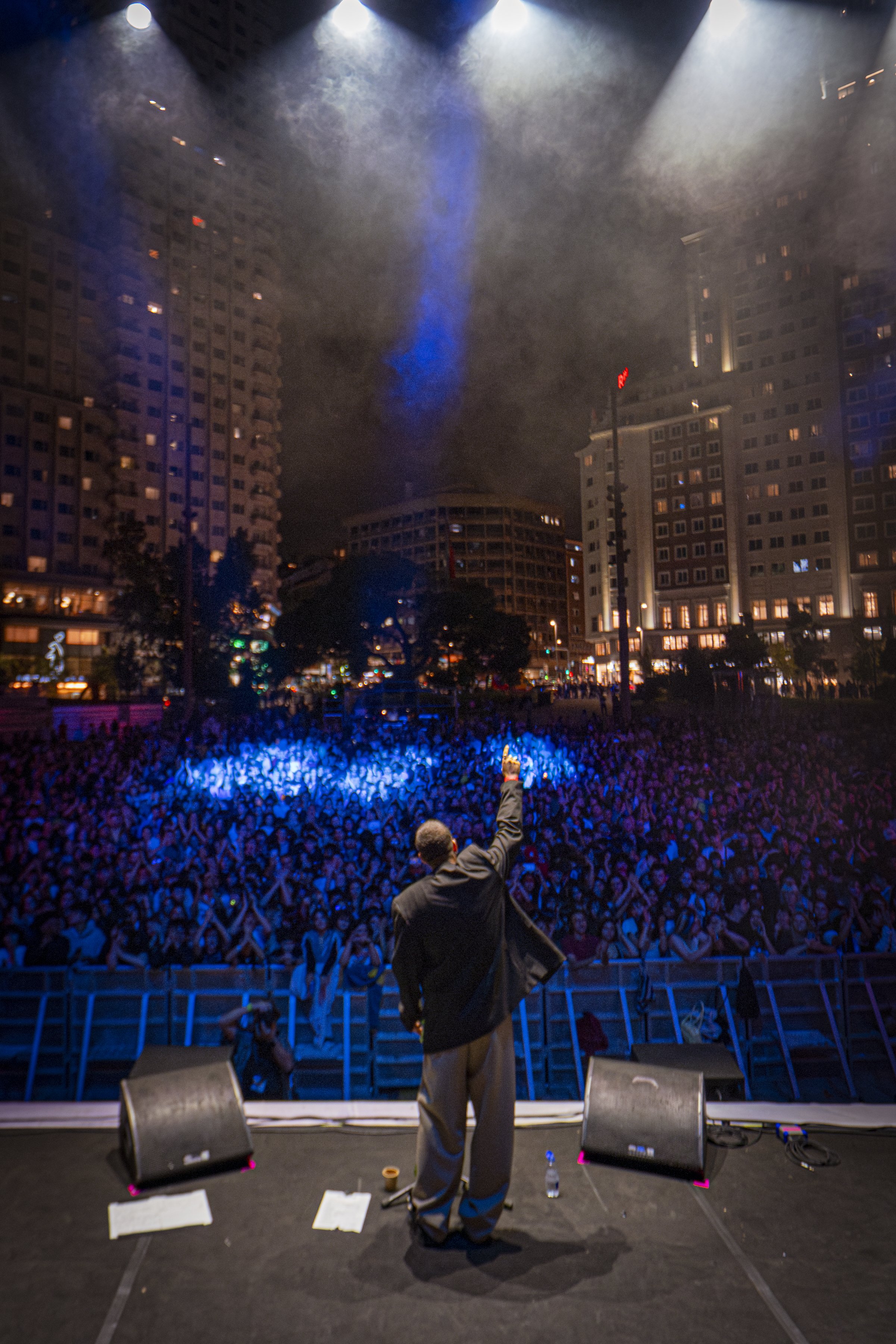 Una persona en el escenario frente a una gran multitud en un concierto en la ciudad, con edificios altos y humo en el cielo.