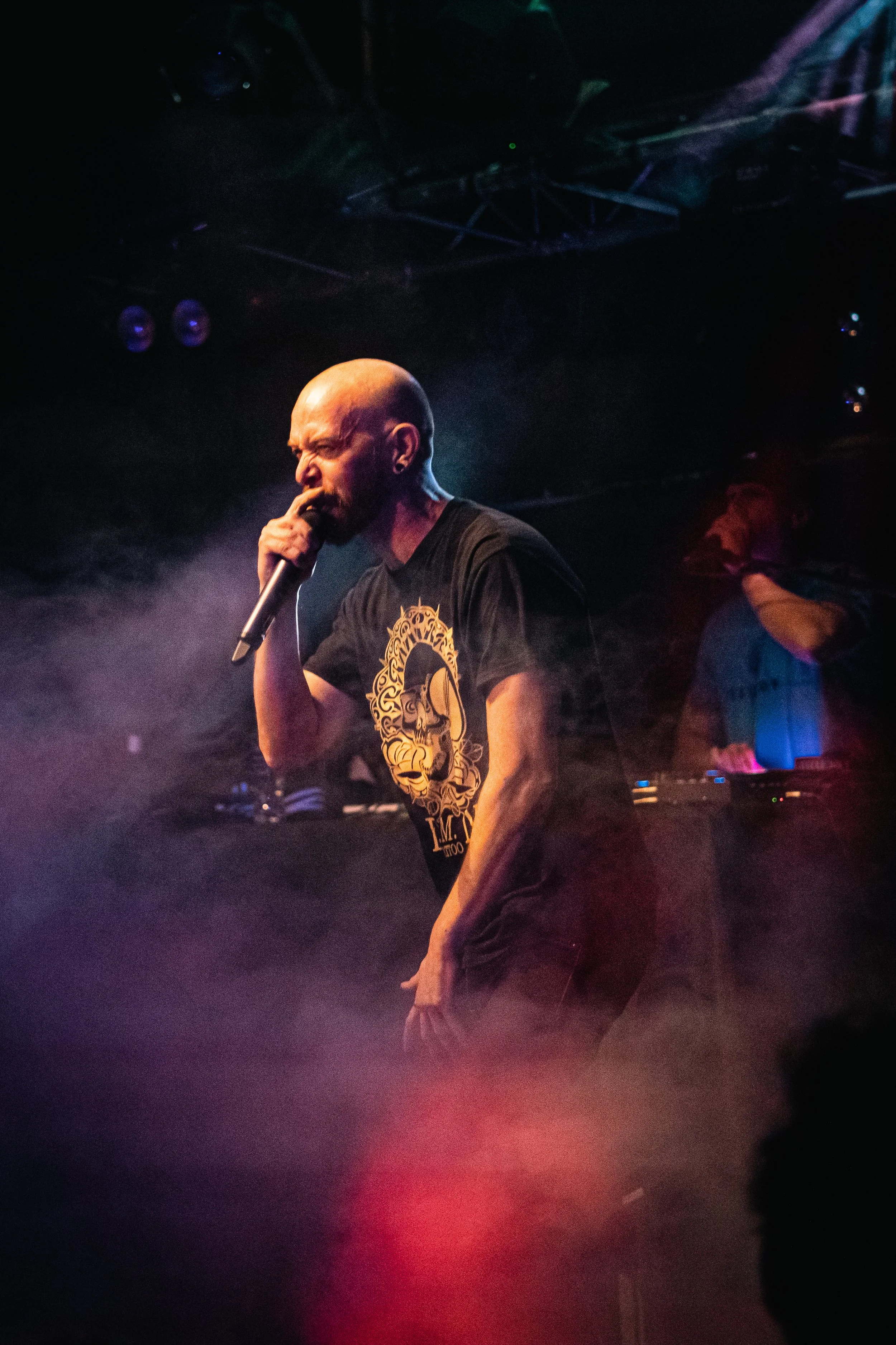 Un hombre con cabeza rapada, sosteniendo un micrófono y cantando en un escenario con iluminación tenue y humo, con otro DJ en el fondo.