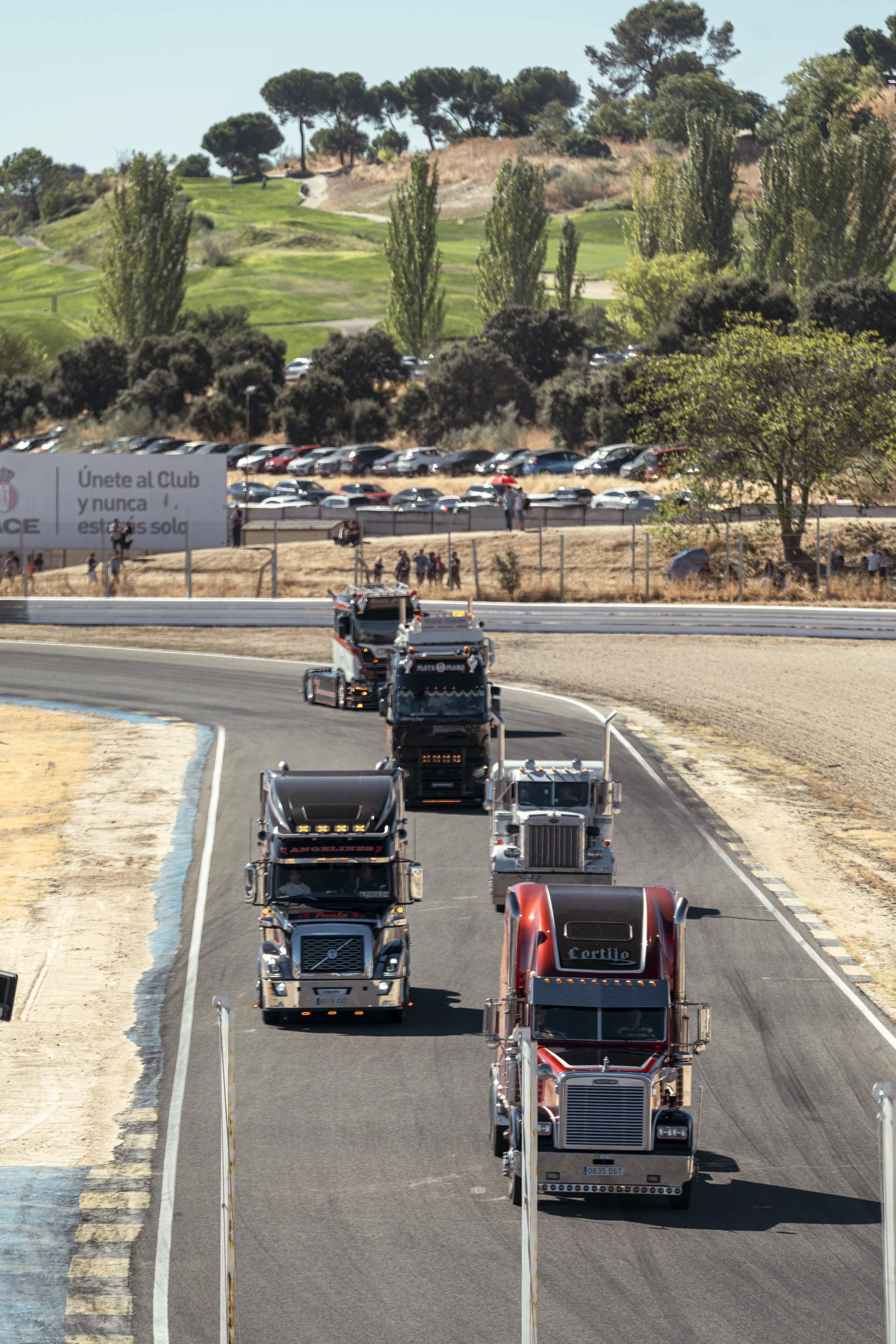 Carril de pista de carreras con varios camiones de carga, en un recorrido de curva. En el fondo hay árboles, un estacionamiento con muchos autos y carteles publicitarios.