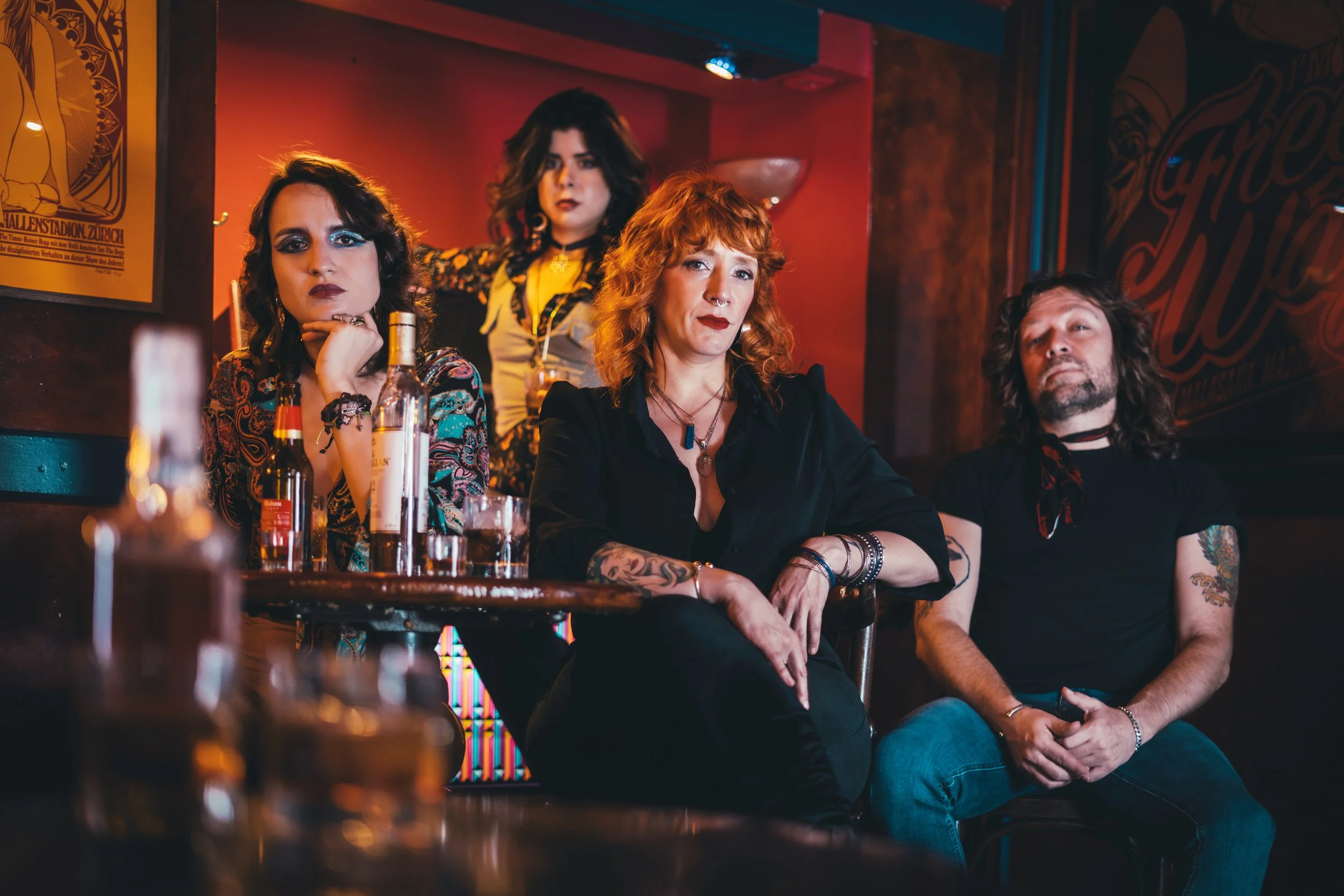 Cuatro personas, tres mujeres y un hombre, sentados en una mesa en un bar con ambiente de rock, con bebidas y música, en un entorno oscuro y colorido.