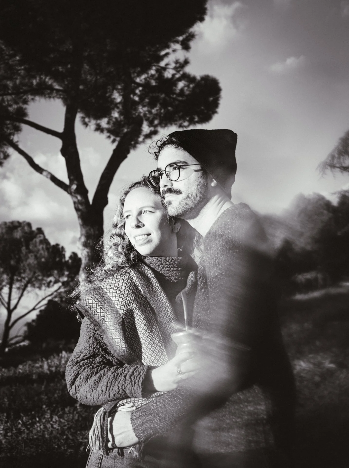 Una pareja abrazada al aire libre con árboles y cielo en el fondo, en blanco y negro.