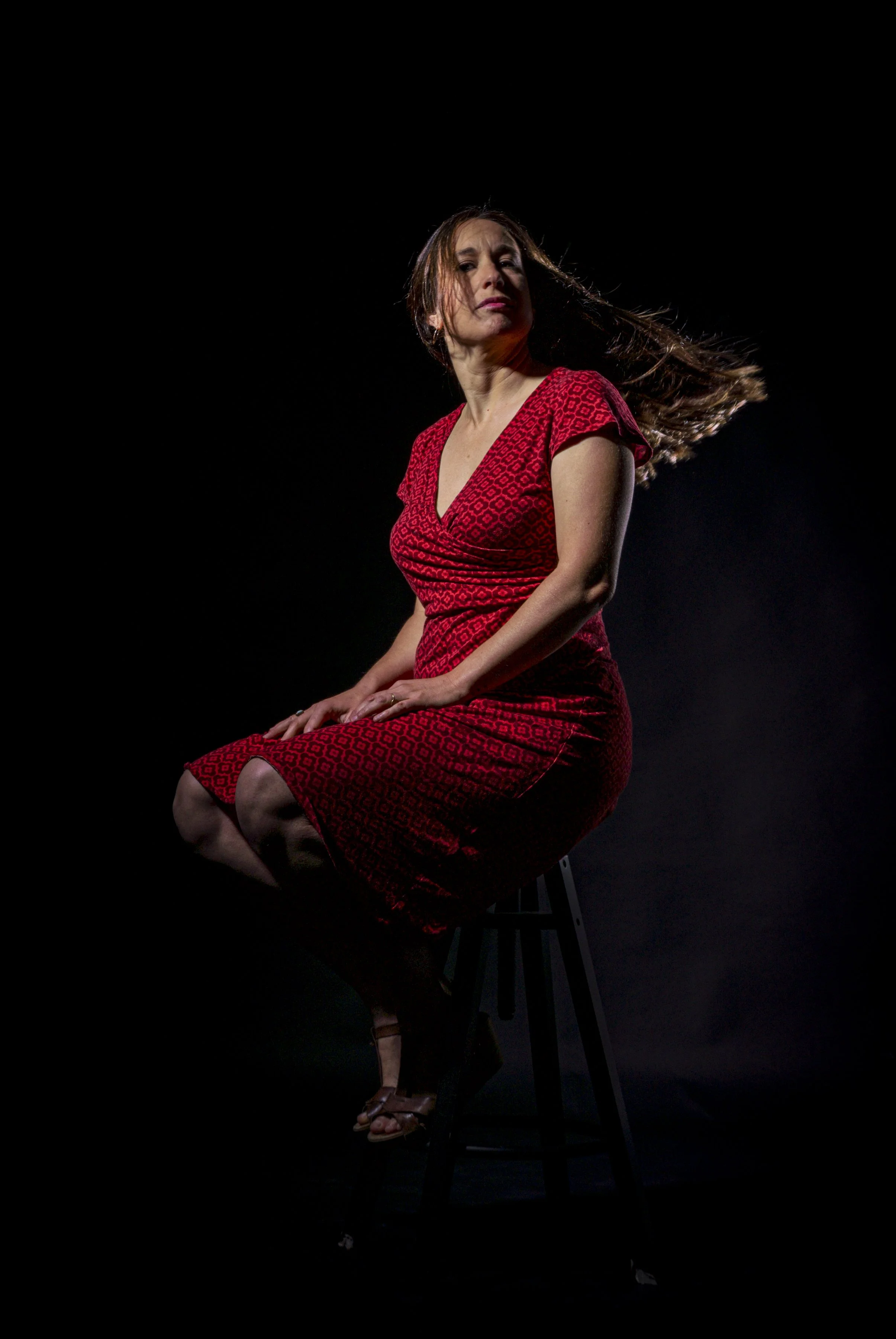 Mujer con vestido rojo sentado sobre un taburete negro en un fondo oscuro, mirando hacia la izquierda y con el cabello movido por el viento.