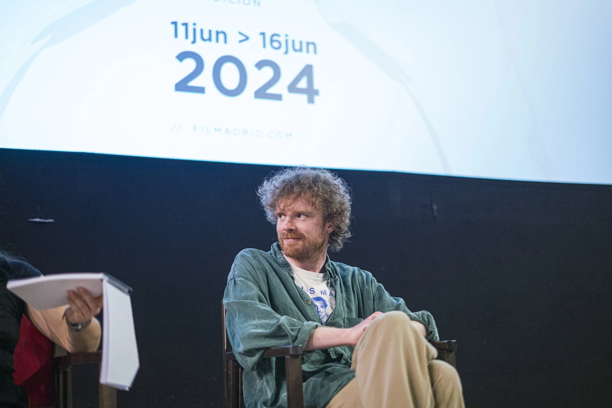 Hombre con barba, cabello rizado y chaqueta verde sentado en una silla en un escenario con pantalla grande de fondo que muestra fechas y texto relacionados con un evento de cine en 2024.