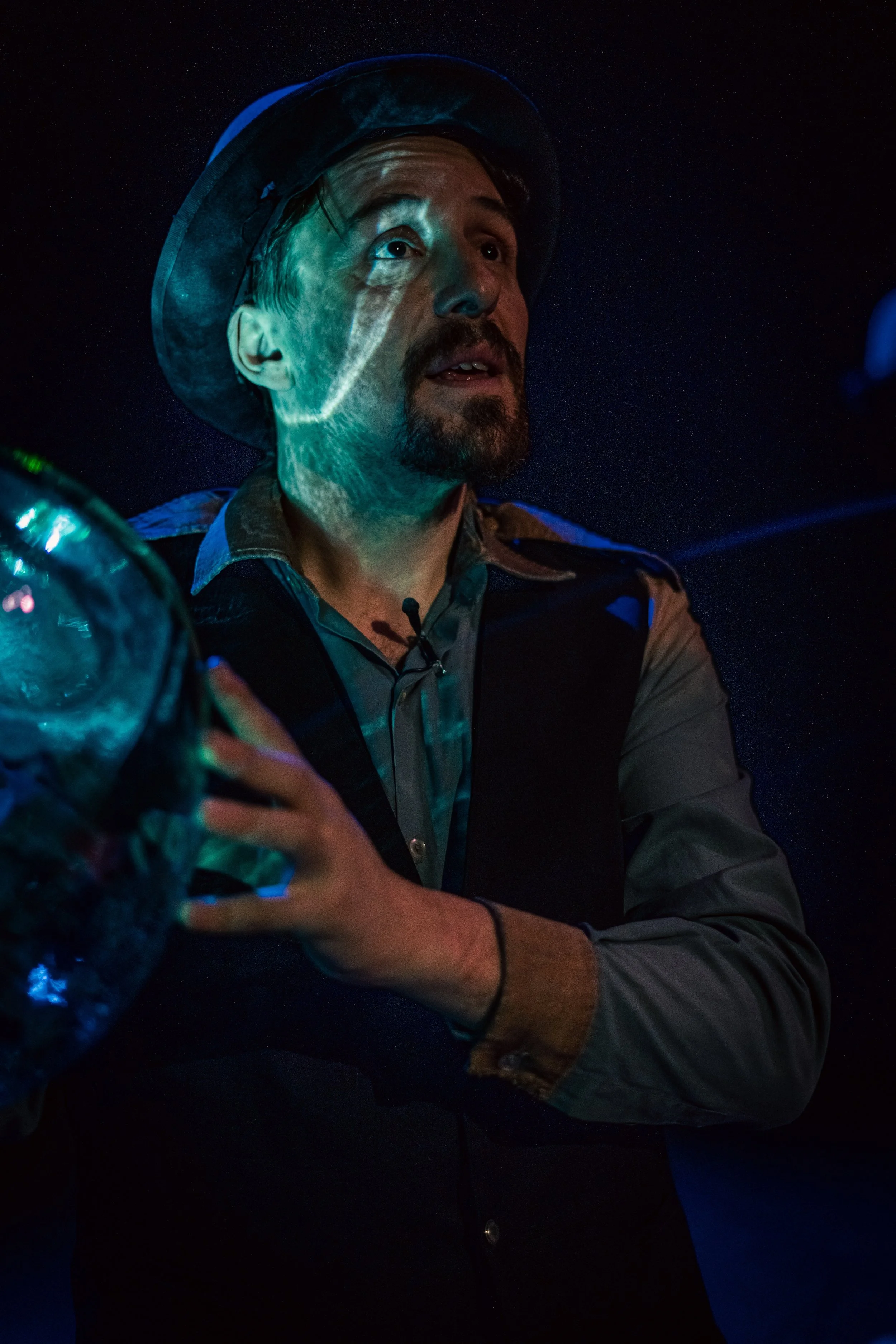 Un hombre con sombrero y barba, sosteniendo un frasco en un ambiente oscuro con iluminación azul y verde, reflejando en el frasco.