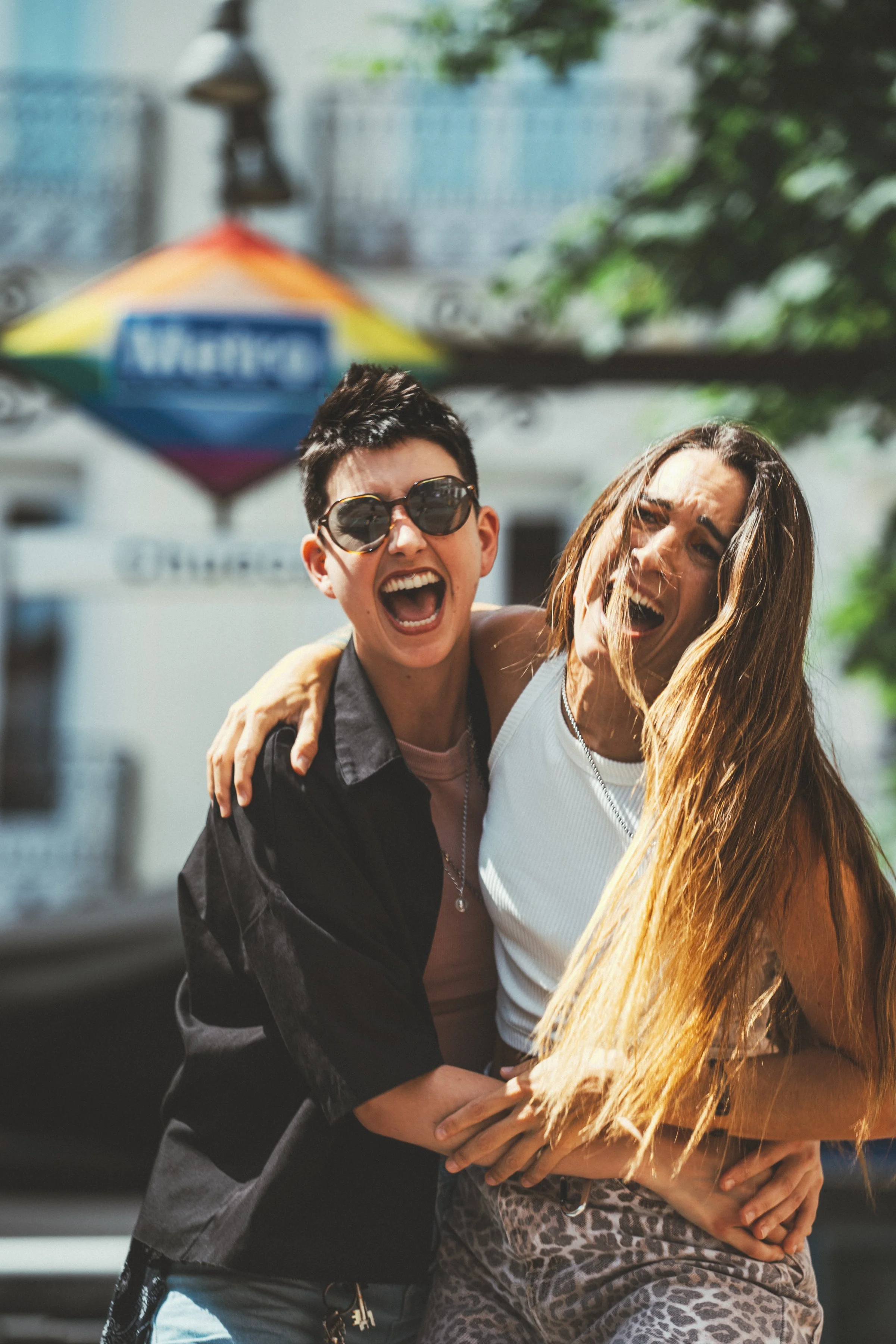 Dos mujeres riendo y abrazadas en la calle durante un día soleado, con un paraguas de colores en el fondo.