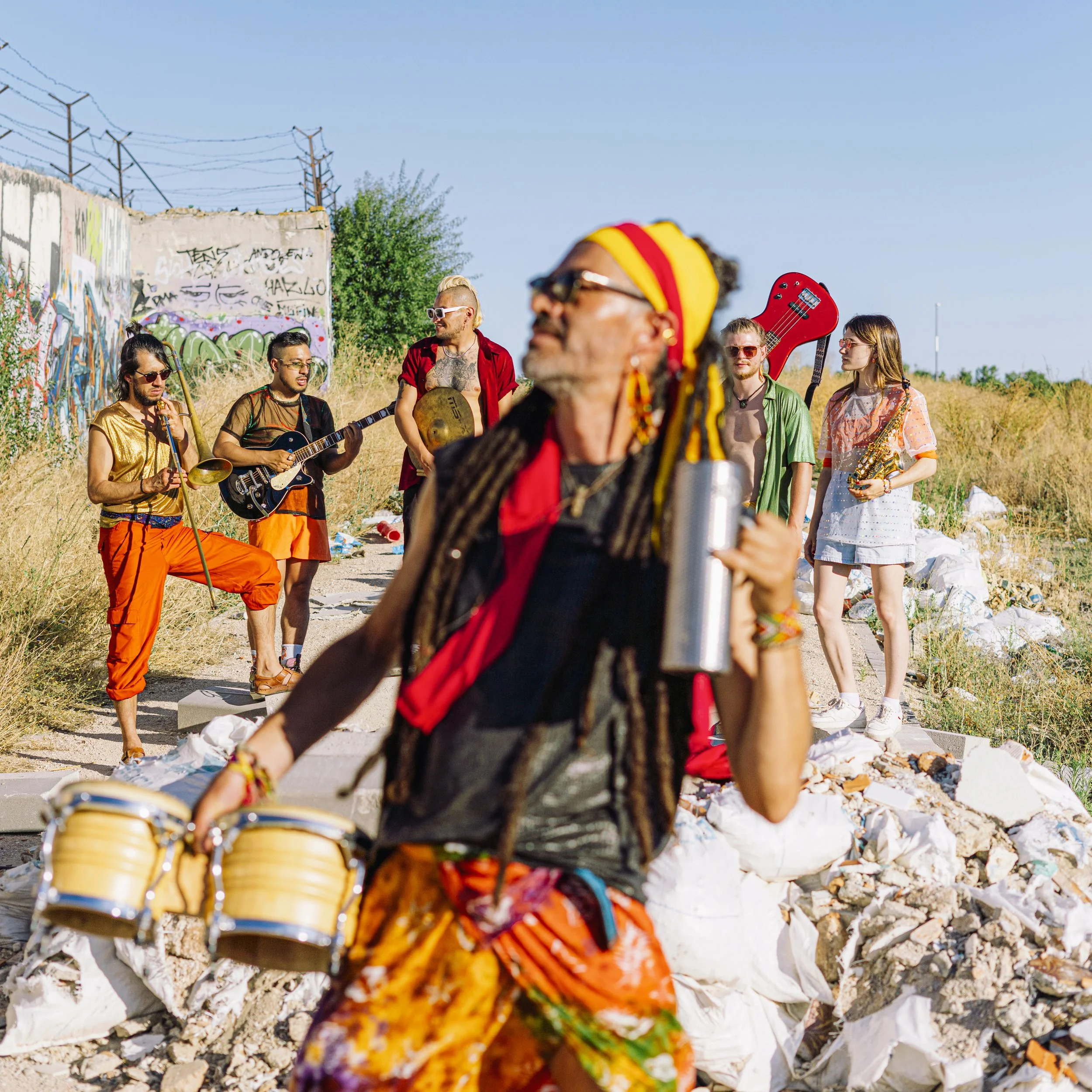 Grupo de músicos en un lugar improvisado, con graffiti en la pared de fondo y rocas alrededor, tocando instrumentos diversos durante el día.