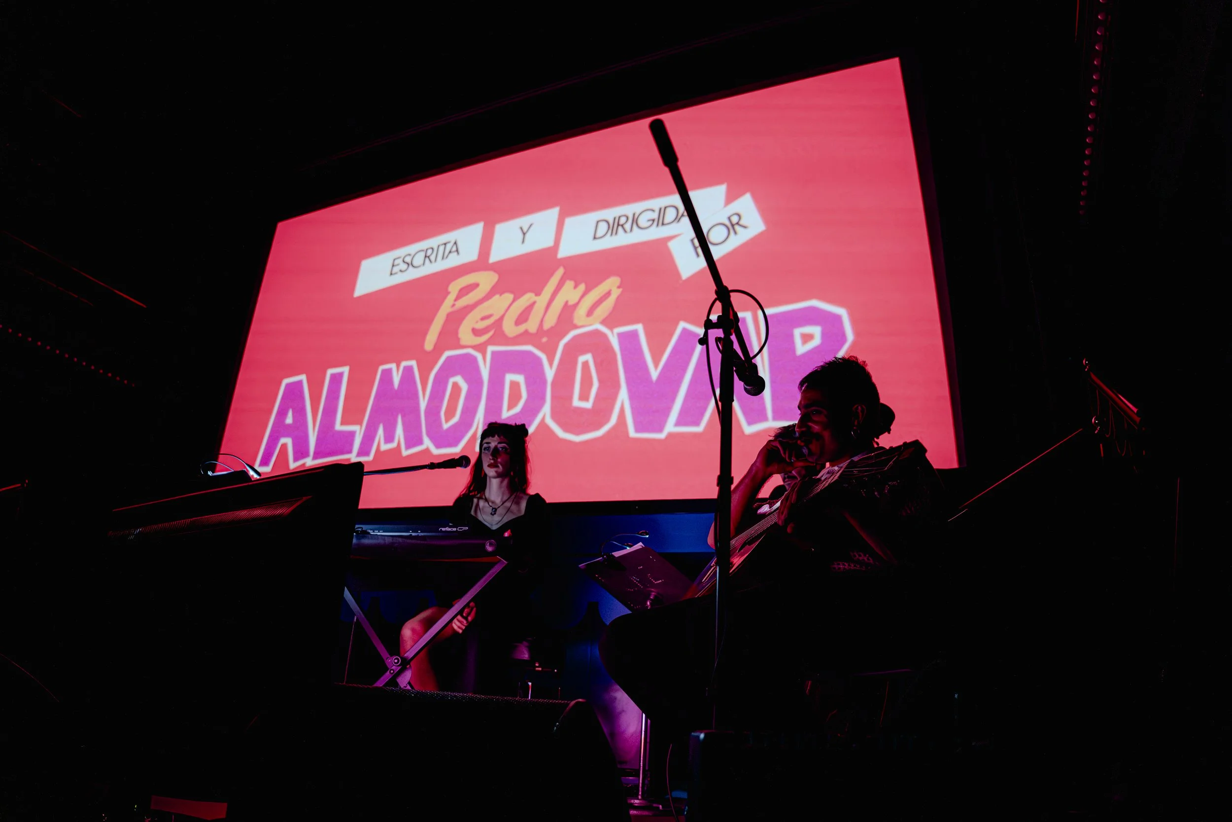 Dos personas en un escenario con pantalla grande al fondo, mostrando el texto en español "Escrita y dirigida por Pedro Almódovar". La persona a la izquierda sostiene una hoja y la de la derecha tiene un micrófono. La escena tiene iluminación tenue co