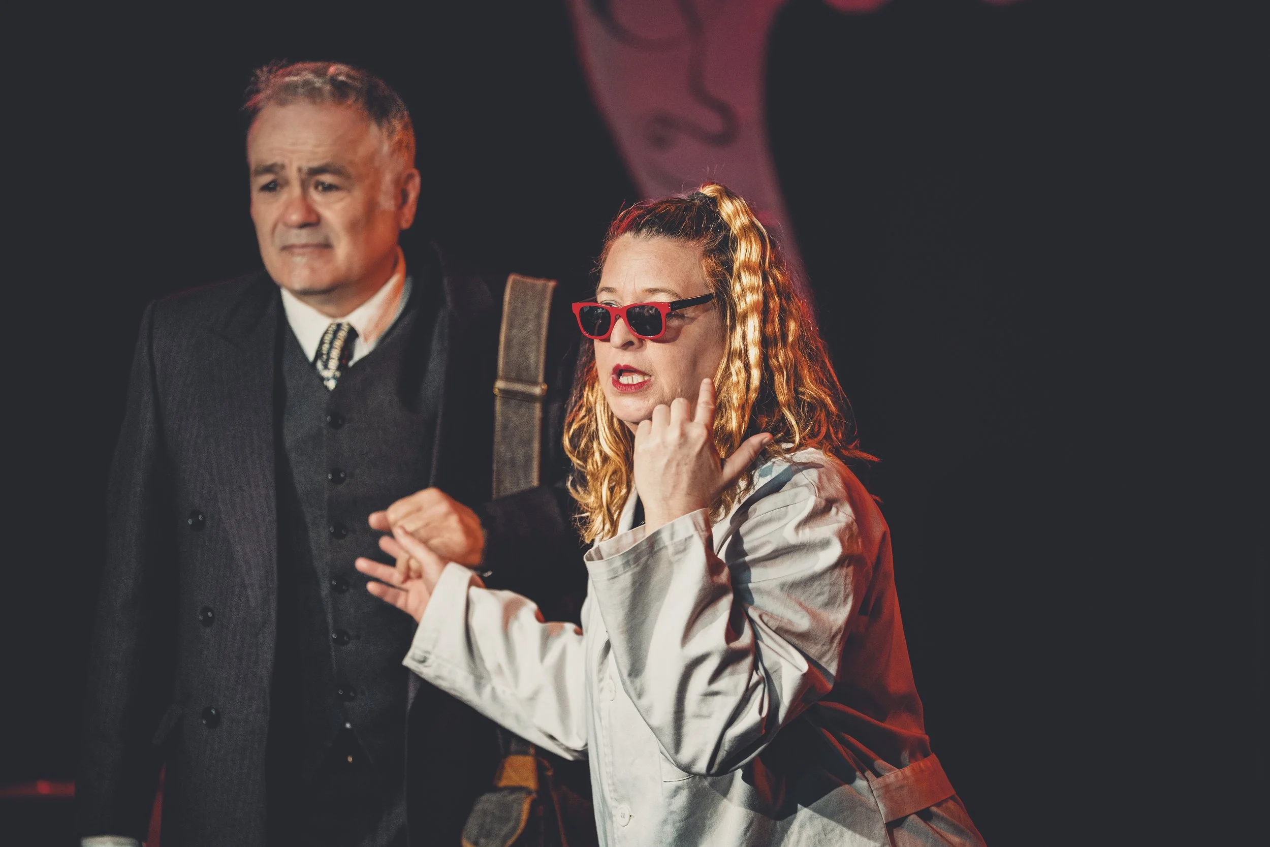 Dos actores en escena, un hombre con vista pensativa y una mujer con gafas rojas, gesticulando dramáticamente.
