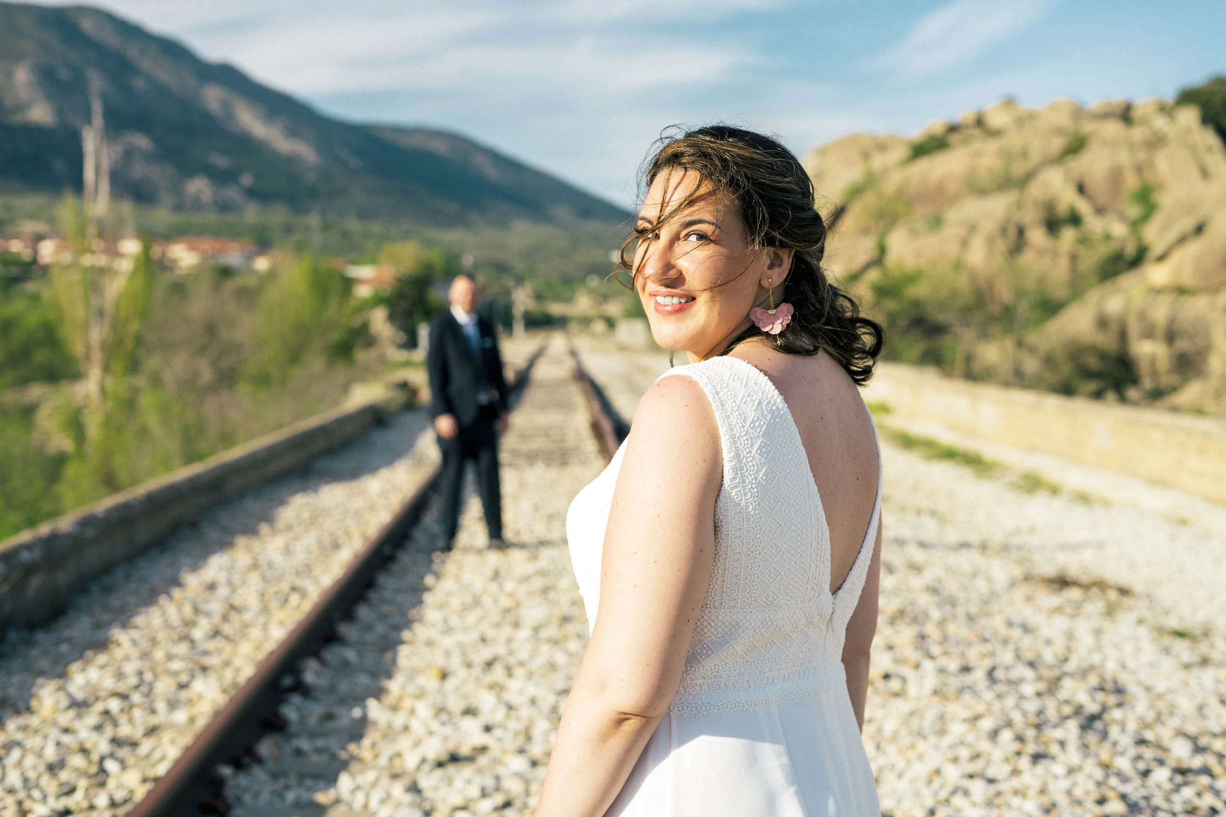 Mujer en vestido blanco con pendientes rosa, sonriendo en una vía de tren con un hombre en el fondo, montañas y vegetación en un paisaje al aire libre.