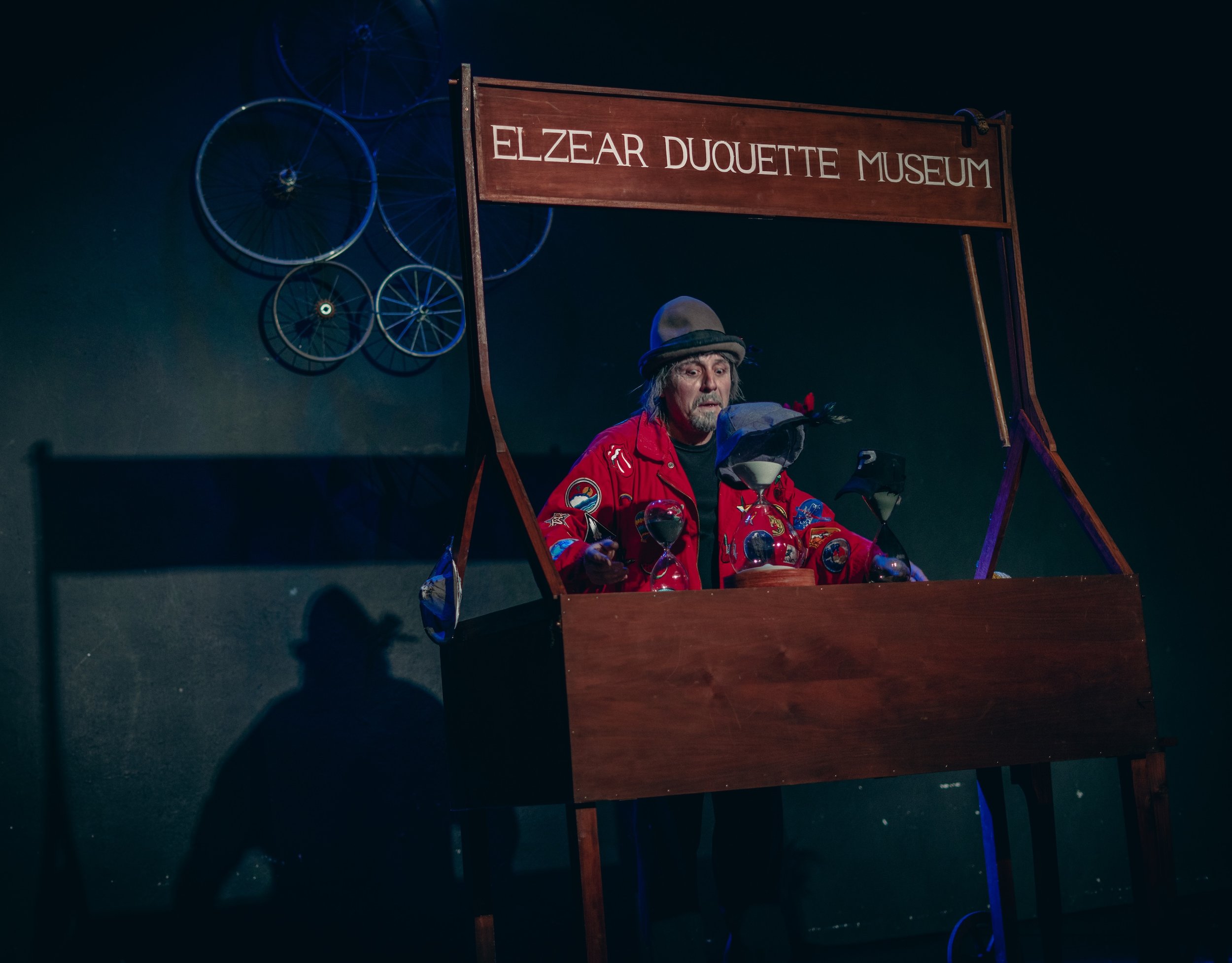 Hombre vestido con chaqueta roja con parches en un escenario con el cartel 'Elzear Duquette Museum', rodeado de aves de papel y bicicletas de metal decorativas en la pared detrás.