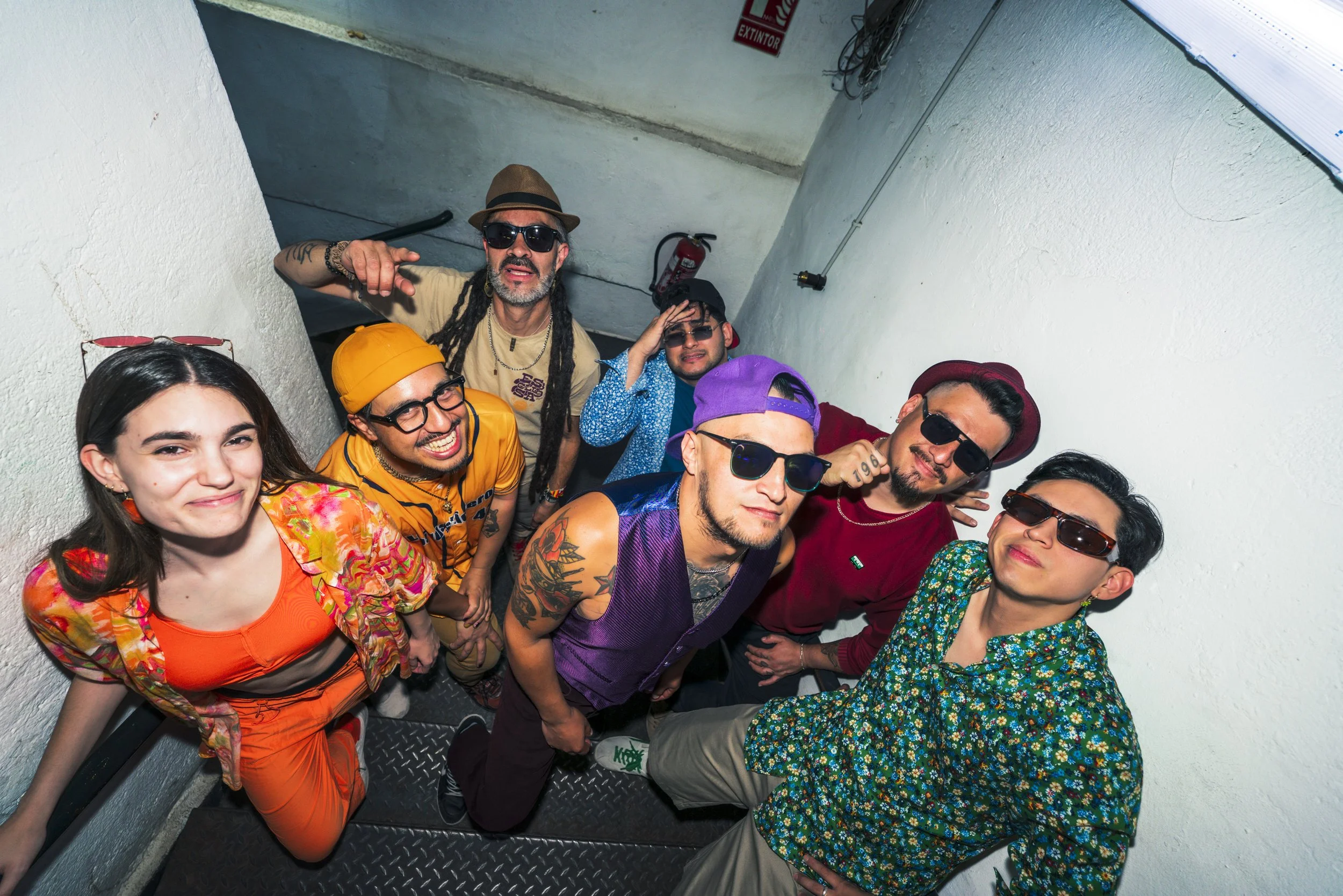 Grupo de personas jóvenes en una escalera, con ropa colorida y accesorios de moda, posando y sonriendo para la cámara en un ambiente informal y alegre.