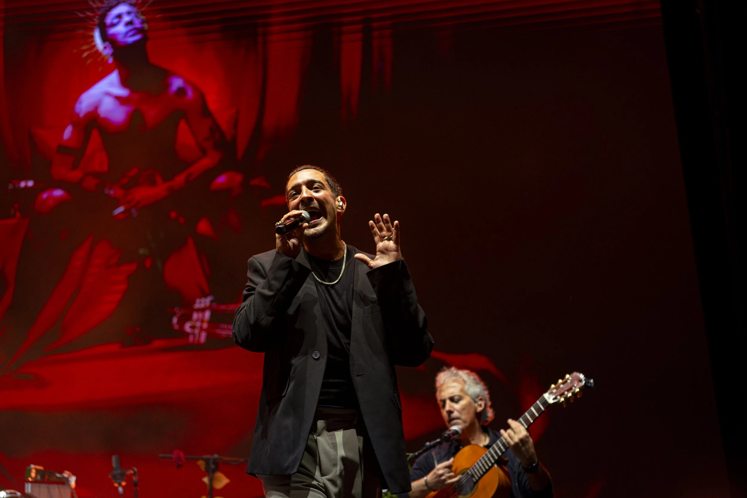 Un cantante de pie en un escenario, sosteniendo un micrófono y con una guitarra, acompañado por otro músico que toca la guitarra. El fondo tiene una imagen grande de un retrato en tonos rojos.