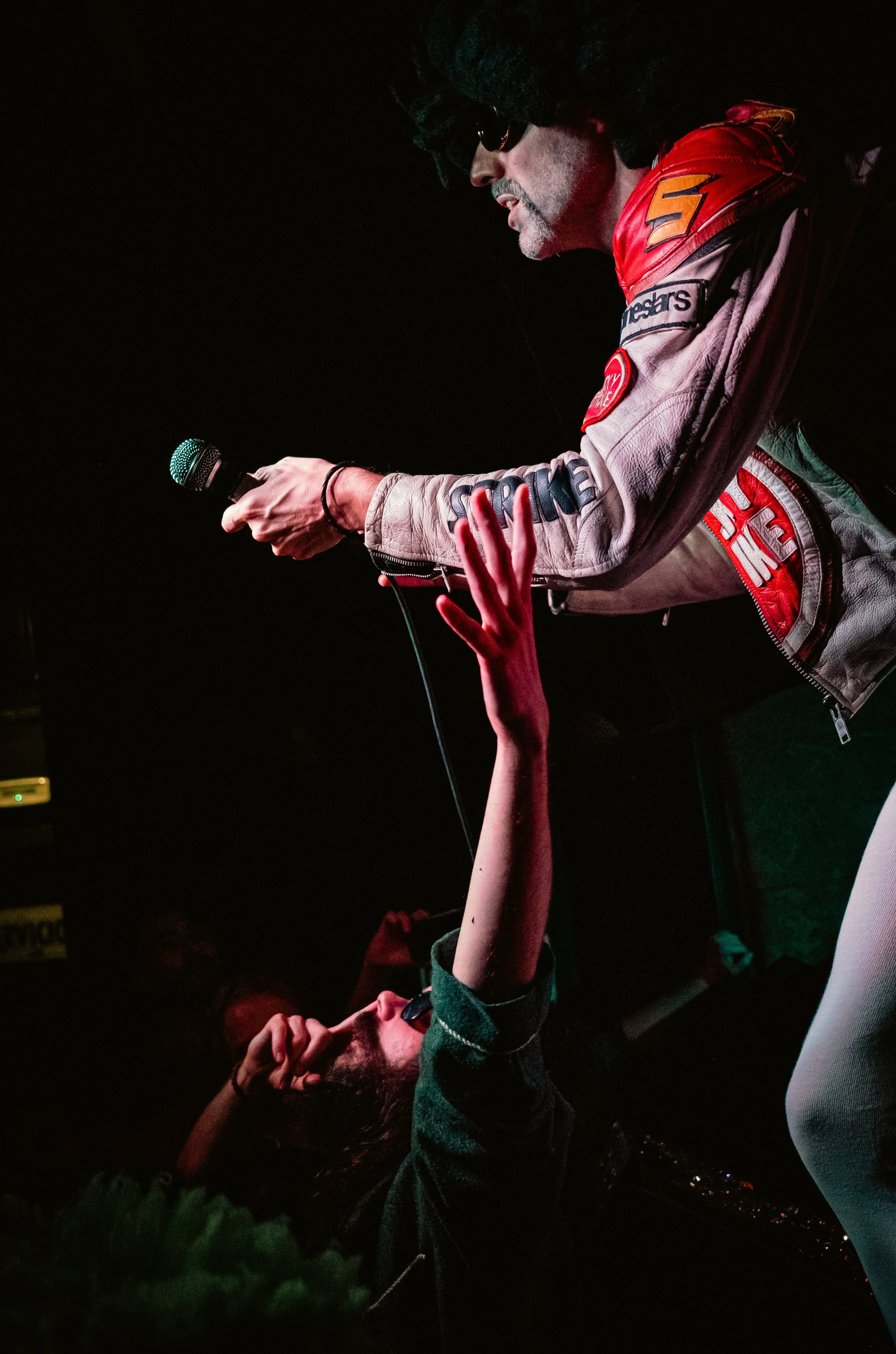 Un hombre con barba, bigote, gafas y un bigote pintado, vestido con una chaqueta con parches, sostiene un micrófono y parece estar en un escenario, mientras una persona en la audiencia le extiende la mano.