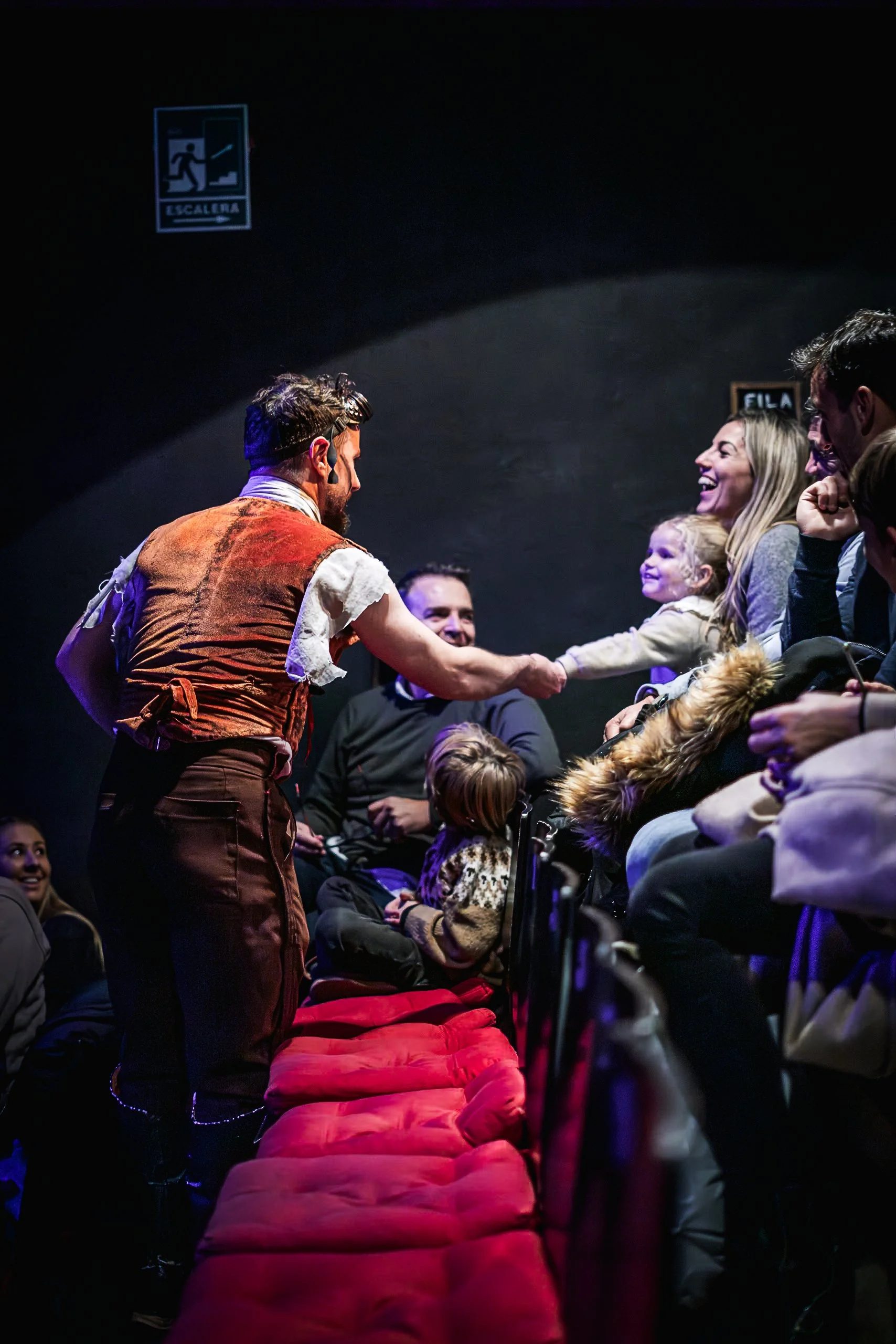 Un hombre vestido con ropa de época interactúa con una niña y otras personas en un teatro, con un fondo oscuro y asientos con cojines rojos.