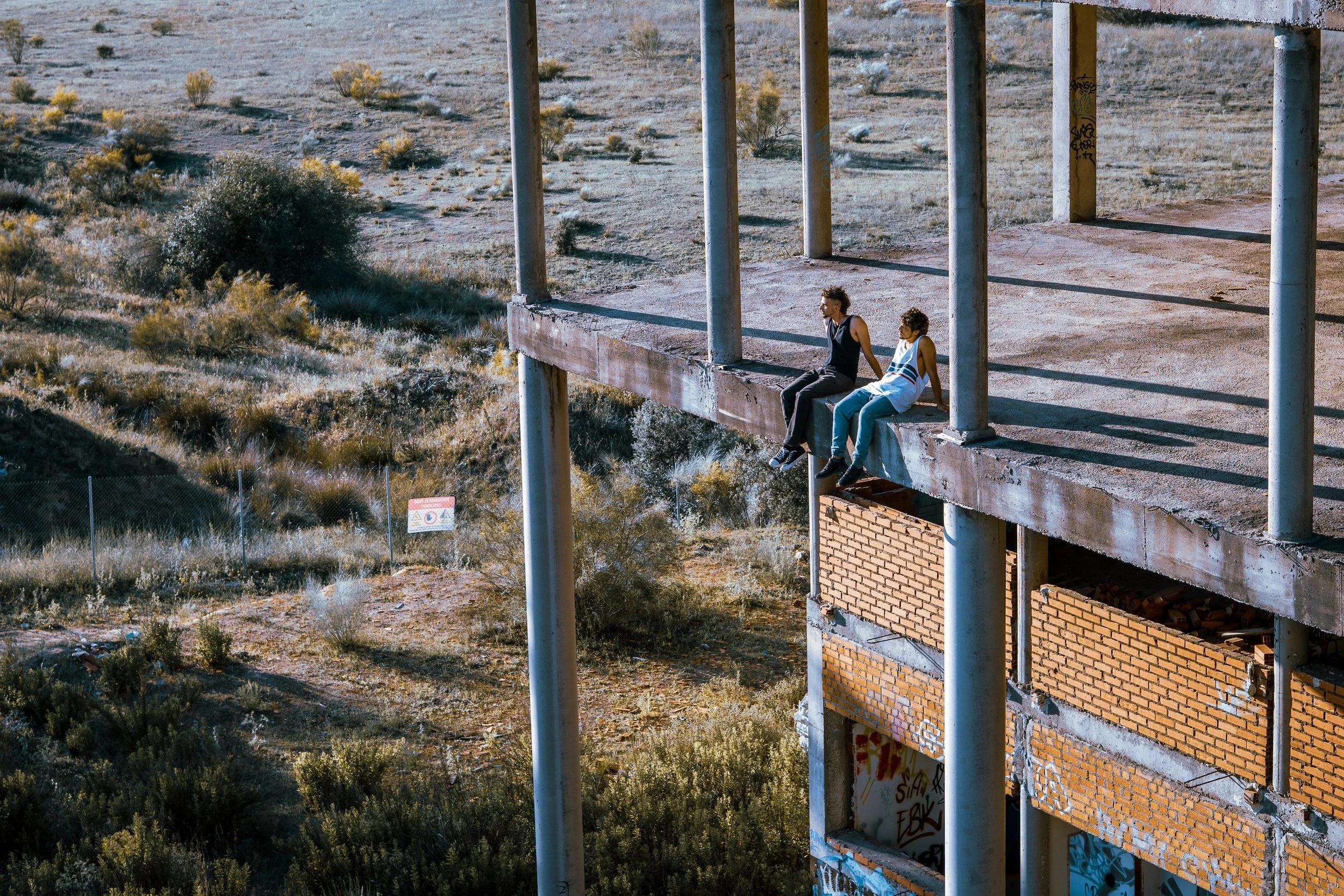 Dos personas sentadas en el borde de una estructura de concreto en un área desértica con vegetación seca, columnas de metal y paredes de ladrillo.
