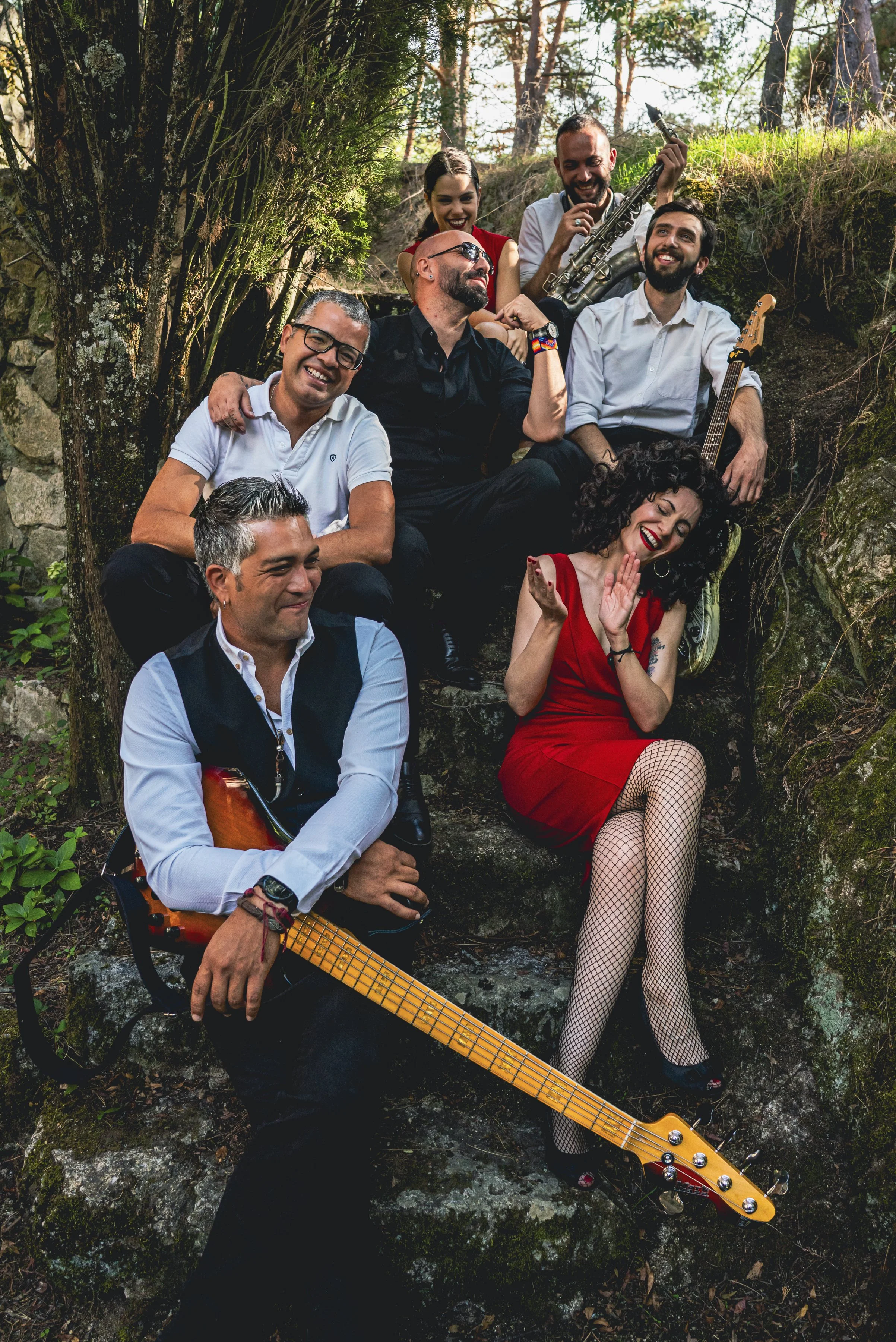 Grupo de ocho personas posando y sonriendo en un entorno natural rodeadas de árboles y rocas, con instrumentos musicales, incluyendo guitarras y saxofón.