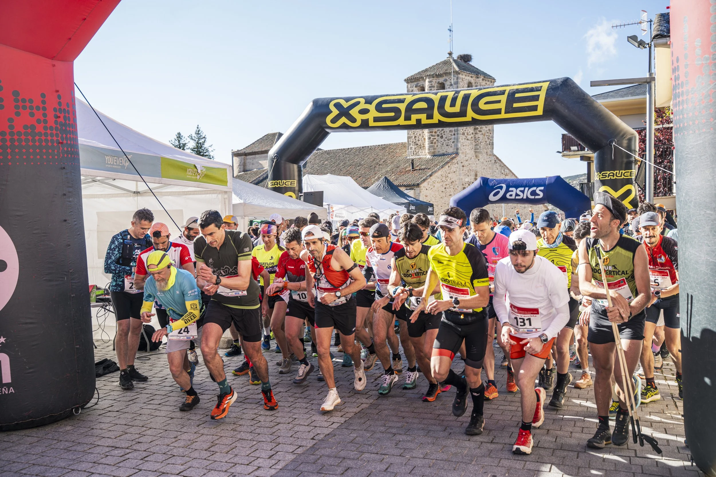 Atletas comenzando una carrera en un evento deportivo al aire libre, bajo arcos de salida con marcas de patrocinadores en un día soleado.