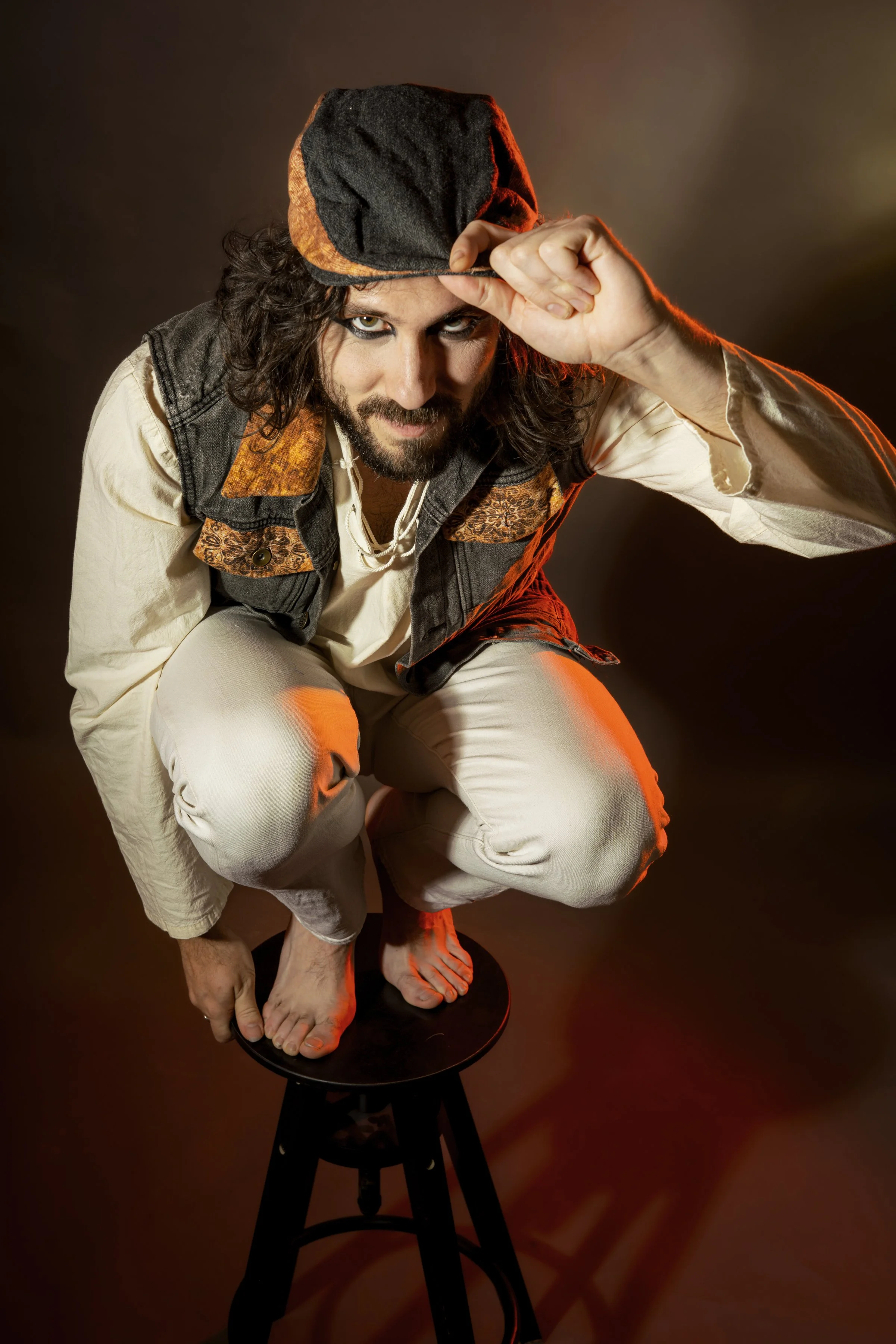 Hombre disfrazado de pirateo, agachado sobre un taburete, con sombrero y ropa de colores oscuros y claros, en un fondo oscuro con iluminación cálida.