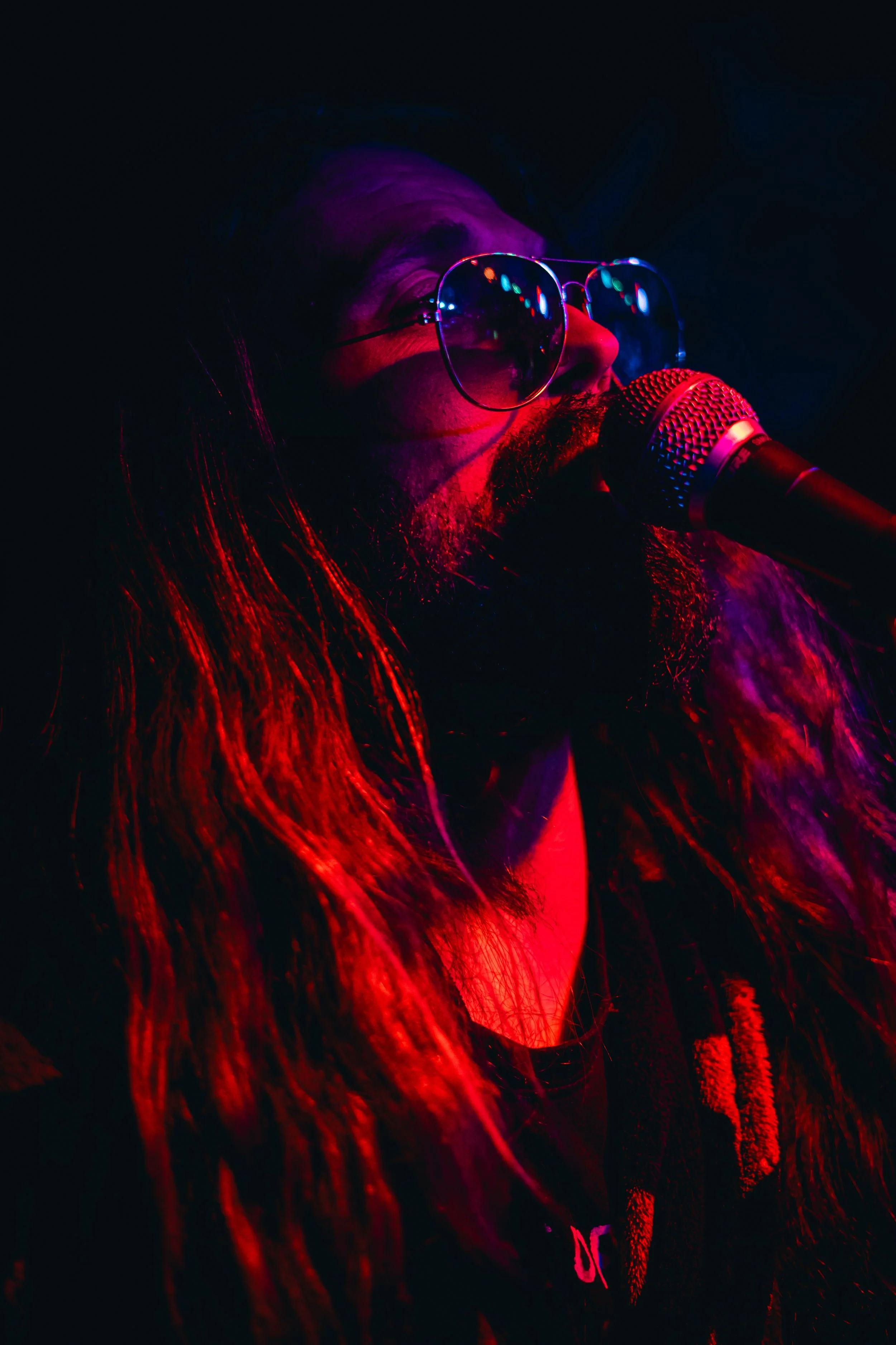 Un hombre con barba y cabello largo, usando gafas de sol, cantando frente a un micrófono en un escenario con iluminación de colores rojos y azules.
