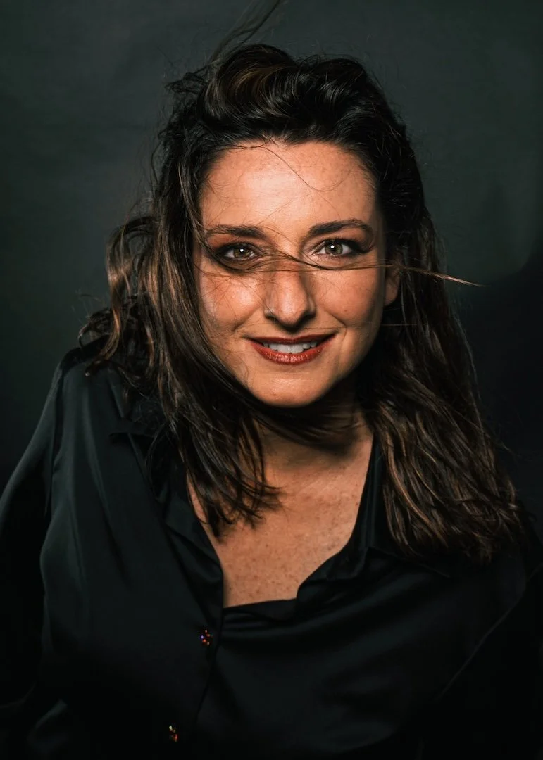 Retrato de una mujer con cabello castaño oscuro y rizado, sonriendo, vestida con blusa negra, fondo oscuro.
