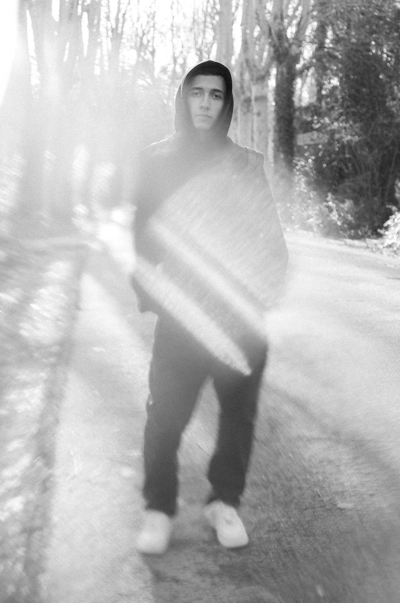 Joven con sudadera con capucha en un camino rodeado de árboles en un día soleado con efectos de luz solar.