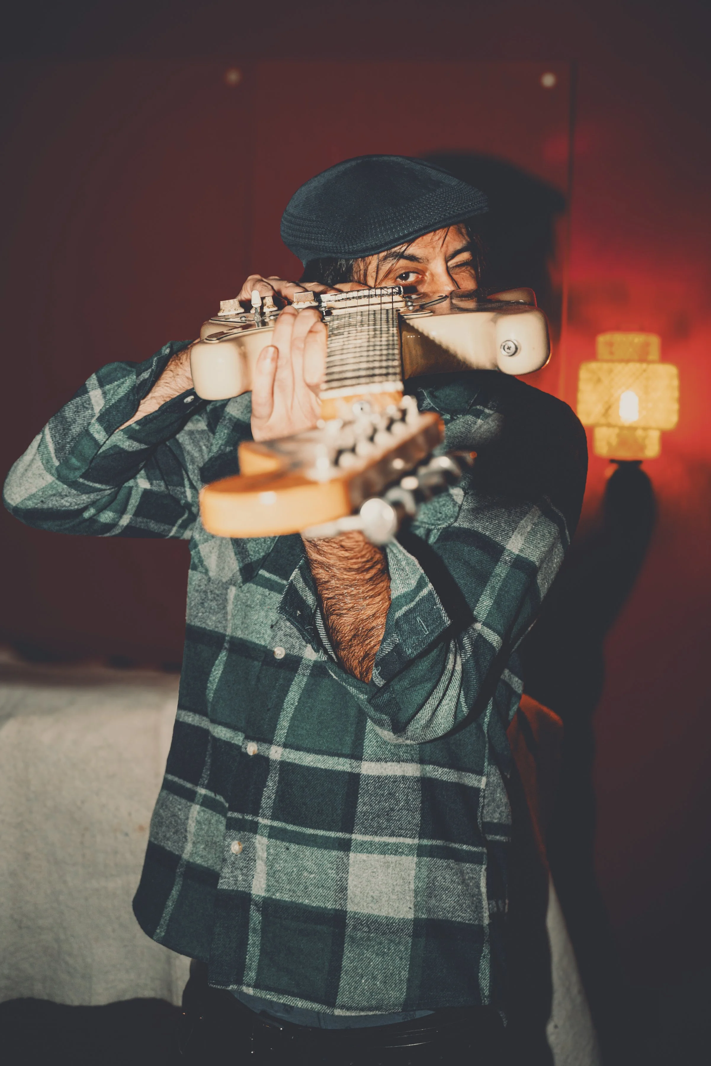 Hombre con gorra y camisa de cuadros sosteniendo una guitarra en posición de disparo, en un ambiente con iluminación cálida.