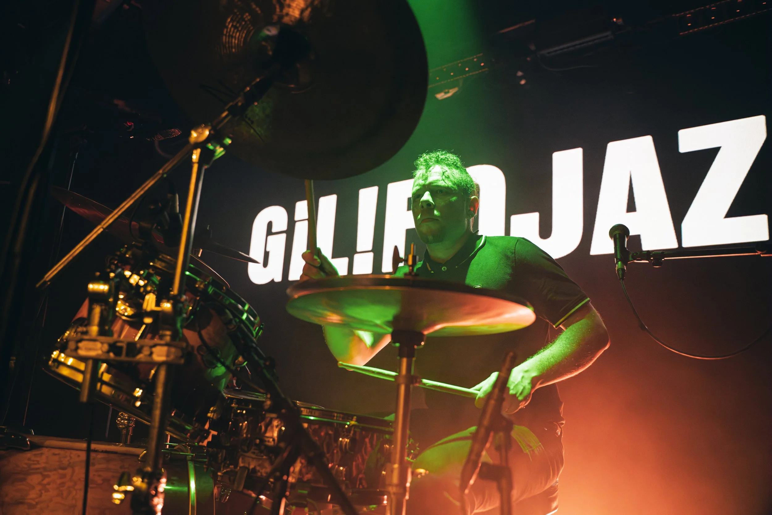 Baterista tocando en un escenario con fondo iluminado que dice 'GILBERTO JAZZ', luz verde y naranja.