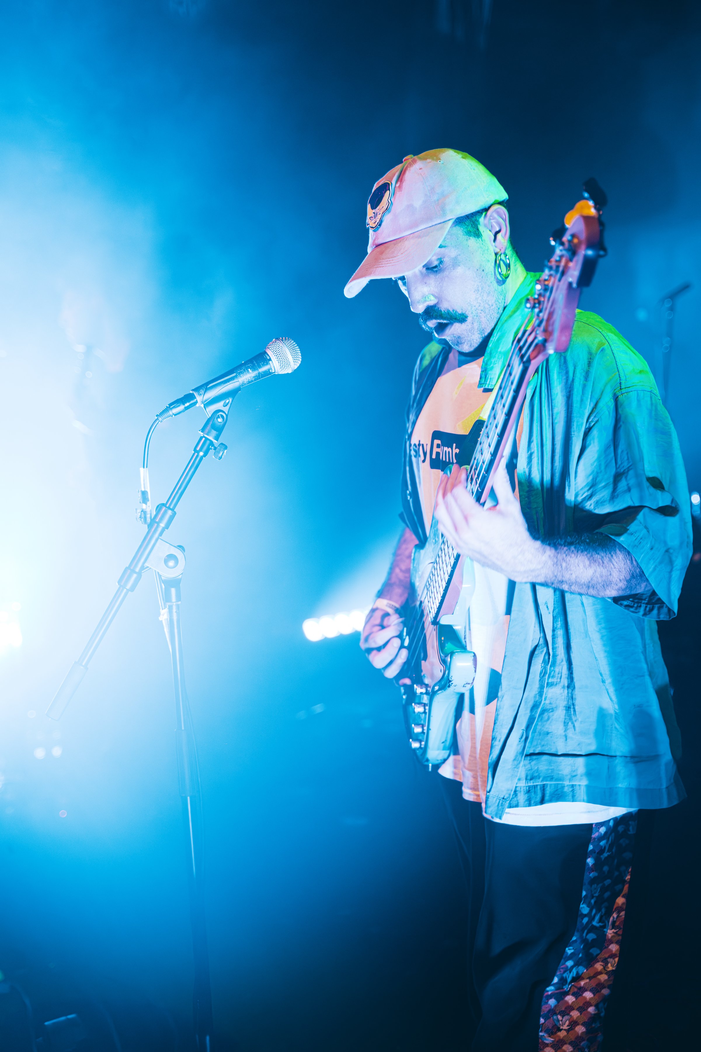 Un hombre tocando la guitarra en un escenario con iluminación azul y verde, con un micrófono frente a él, y lleva una gorra y ropa casual.