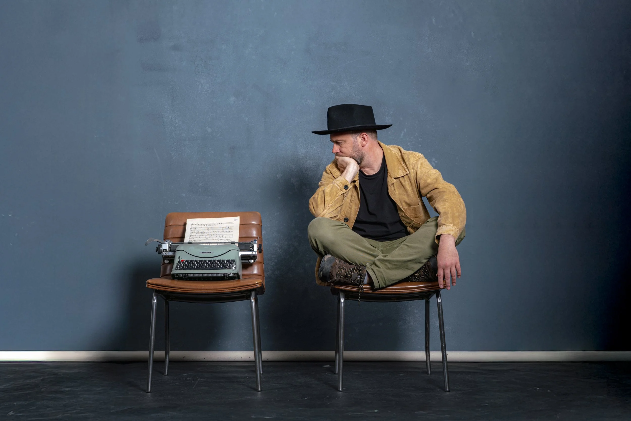 Hombre con sombrero negro, chaqueta marrón y pantalones verde oliva, sentado en una silla junto a una máquina de escribir, en una pared azul.