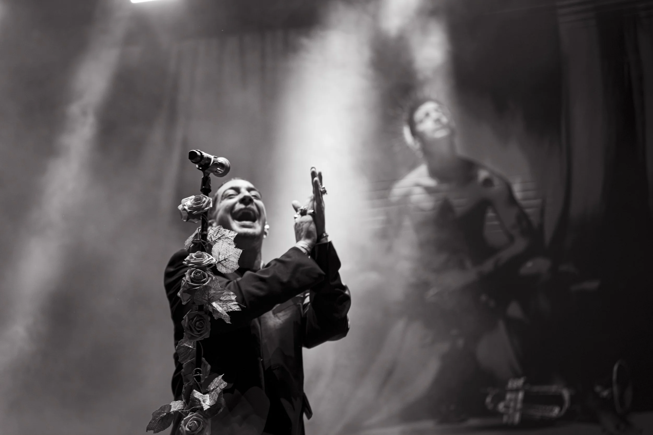 Una mujer canta apasionadamente en un escenario con un micrófono decorado con rosas, mientras que en el fondo se refleja su rostro en un espejo en una imagen en blanco y negro.