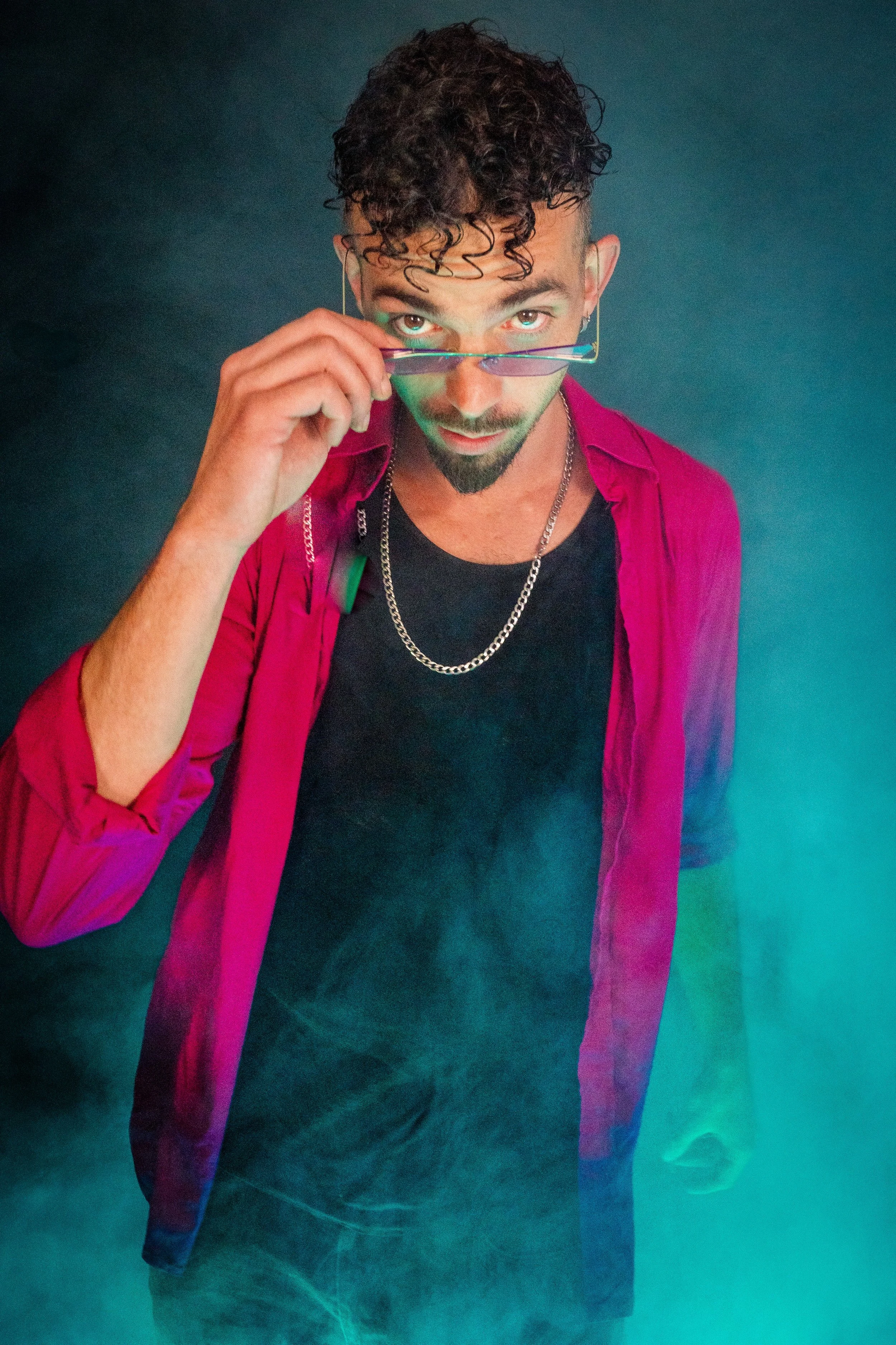 Hombre con cabello rizado y barba, usando gafas y ropa de colores vibrantes, en un fondo oscuro con humo o niebla de color verde azulado.
