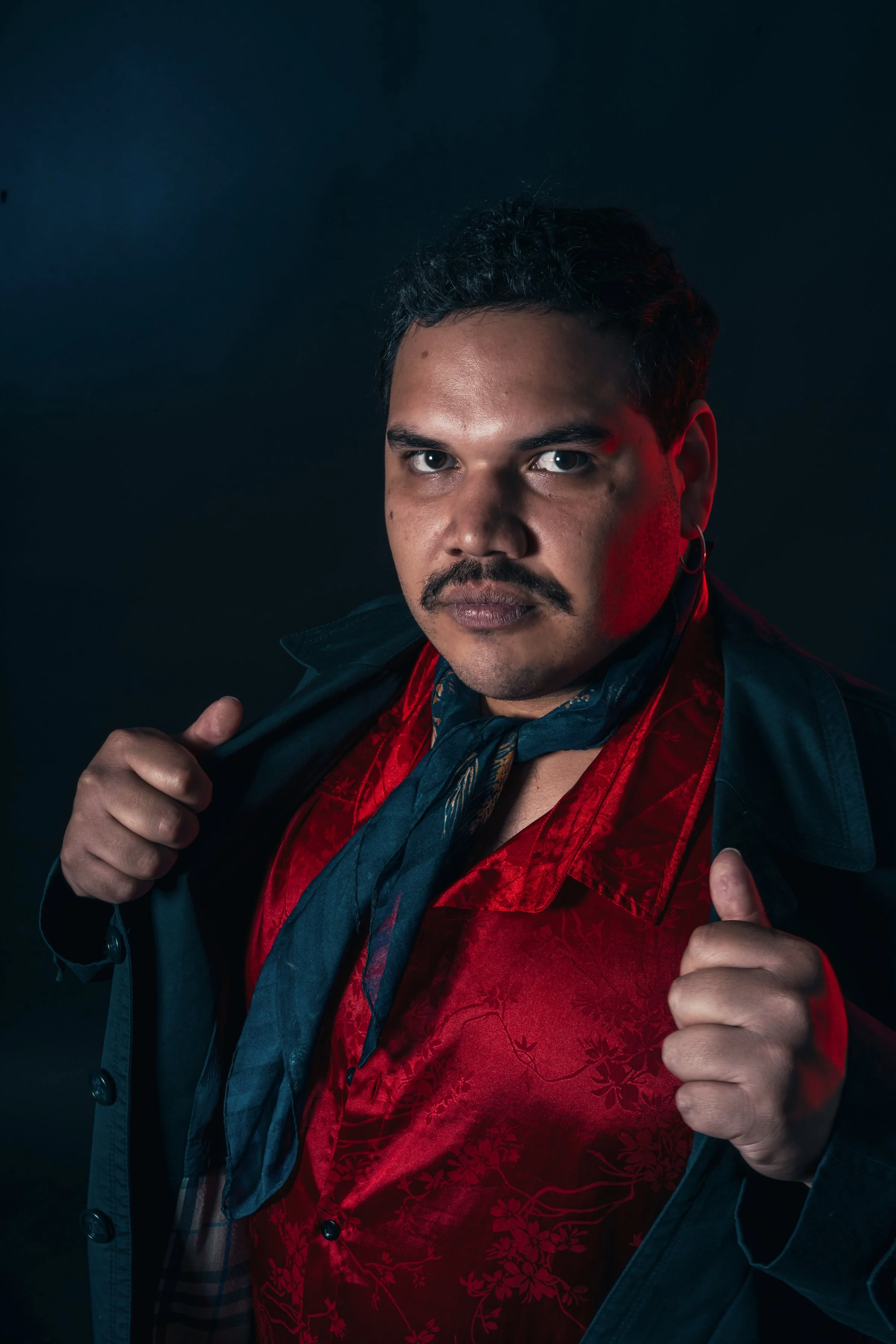 Hombre con bigote y barba, vestimenta llamativa con chaqueta negra, camisa roja con diseño floral y bufanda azul, posando en un fondo oscuro, con expresión segura y desafiante.