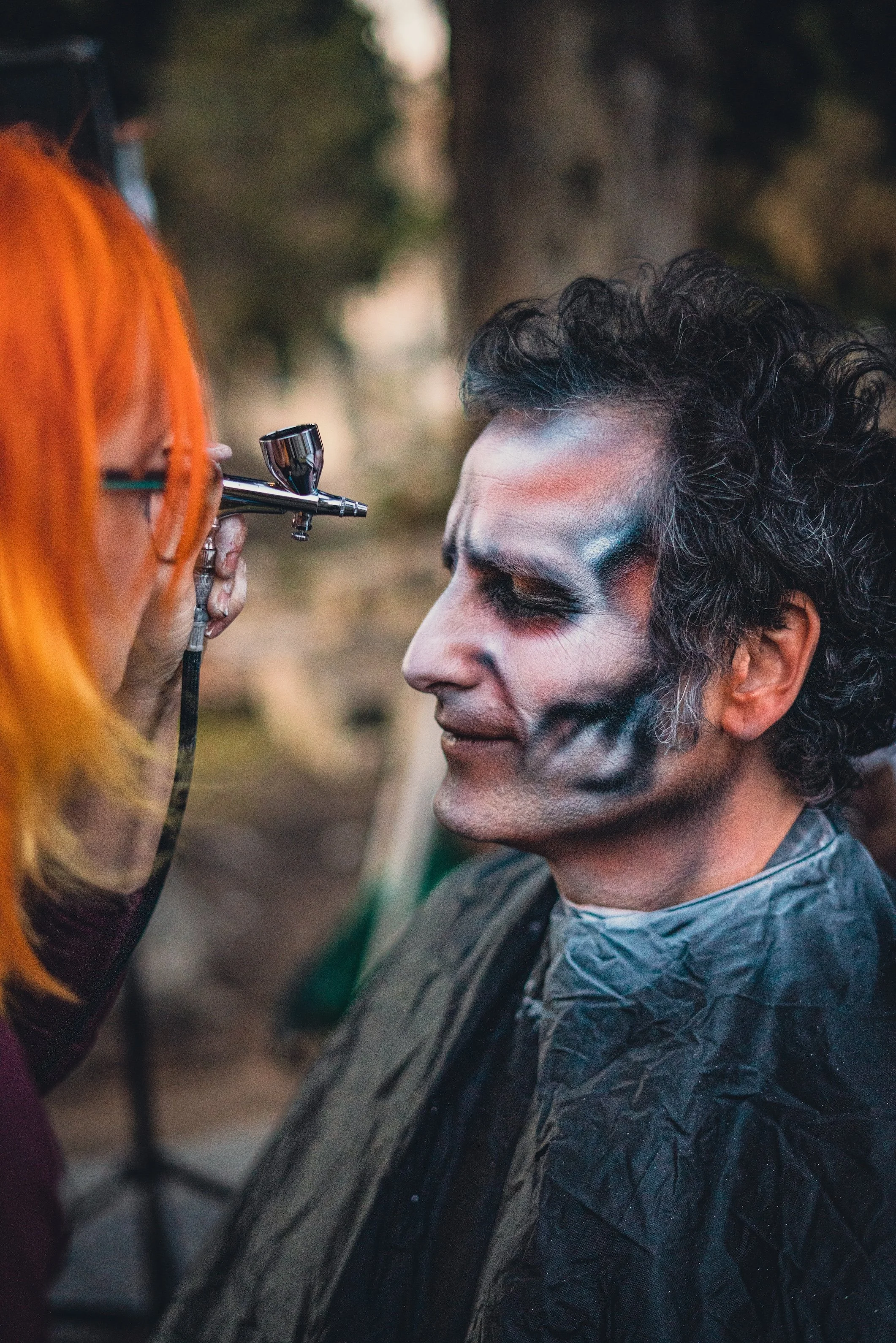 Alguien se está haciendo maquillaje de efectos especiales en el rostro de un hombre con piel clara y cabello rizado, en un entorno al aire libre.