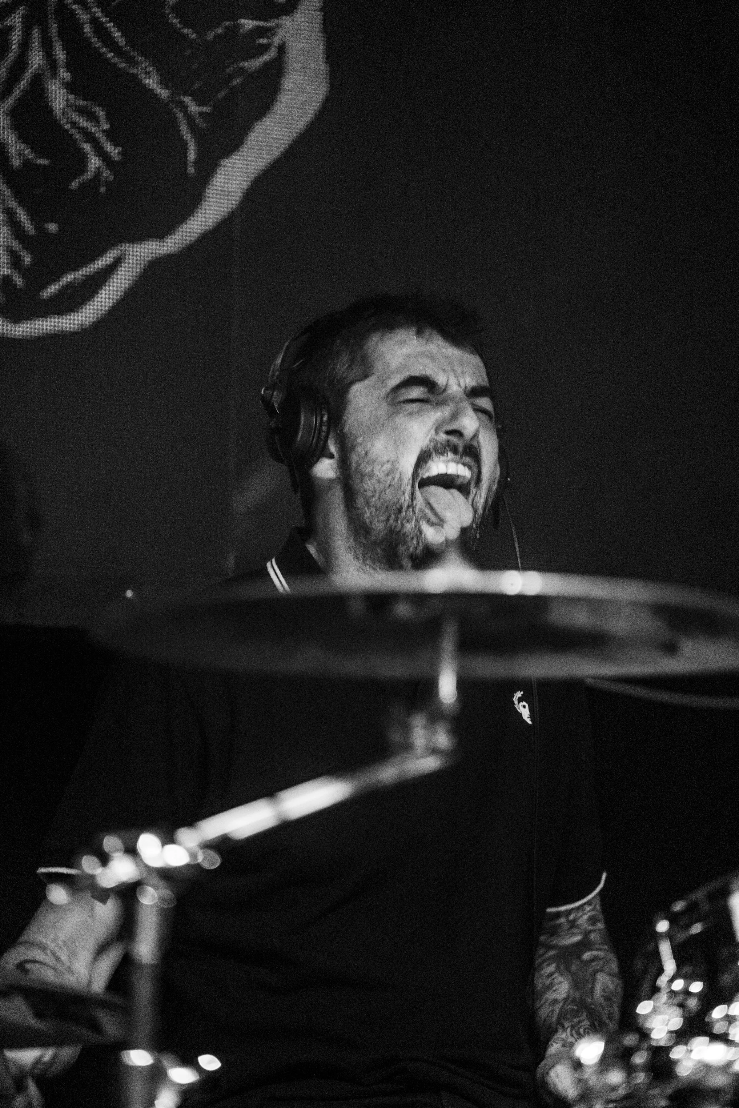 Un hombre tocando la batería con expresión apasionada, usando auriculares, en un escenario oscuro en blanco y negro.