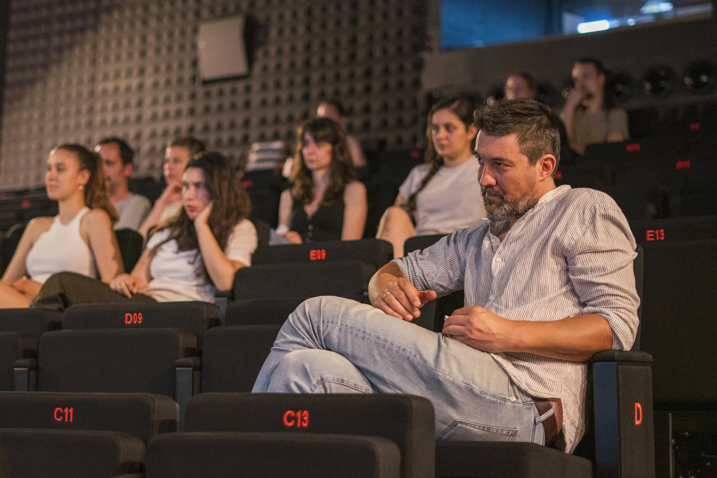 varias personas sentadas en un auditorio, algunas con atención y otras con expresión pensativa, en un ambiente de teatro o sala de conferencias