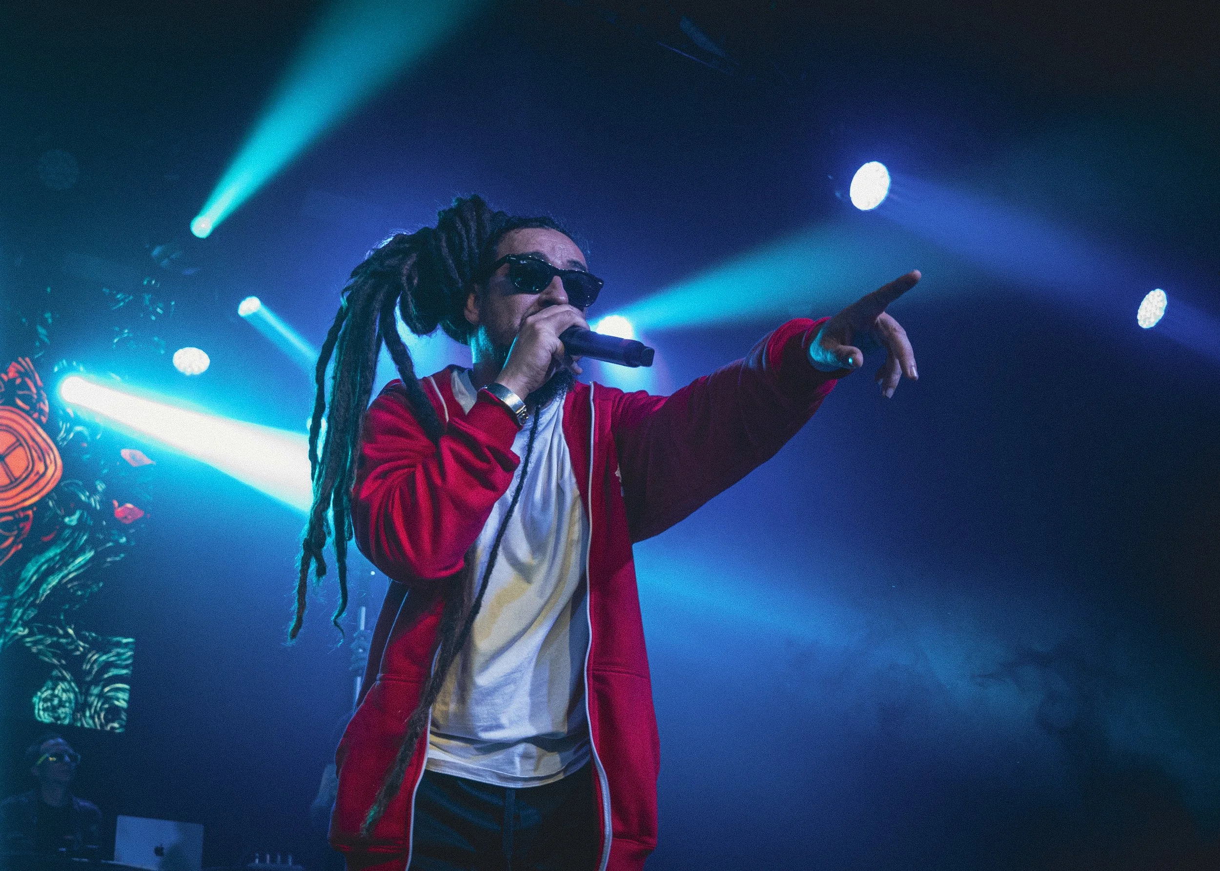 Un cantante con dreadlocks, gafas negras y chaqueta roja actuando en un escenario con luces azules y blancas, señalando con una mano mientras sostiene un micrófono con la otra.