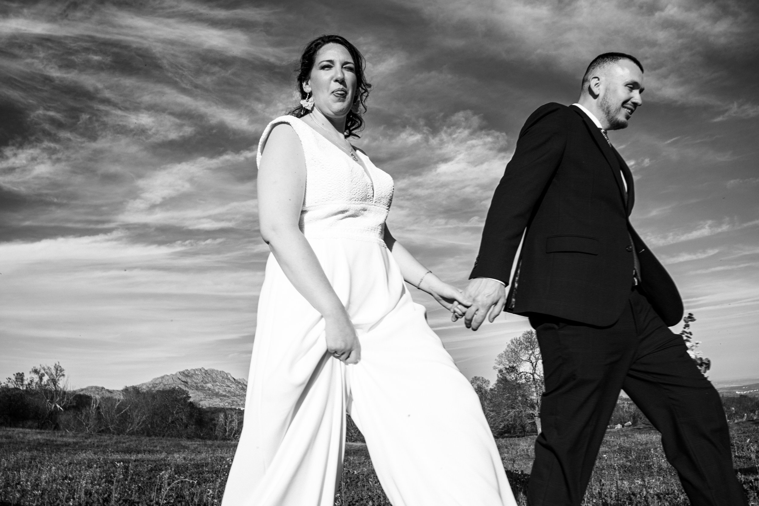 Una pareja vestida con ropa formal caminando en un campo con un cielo nublado al fondo, la mujer lleva un vestido blanco y el hombre lleva un traje negro.