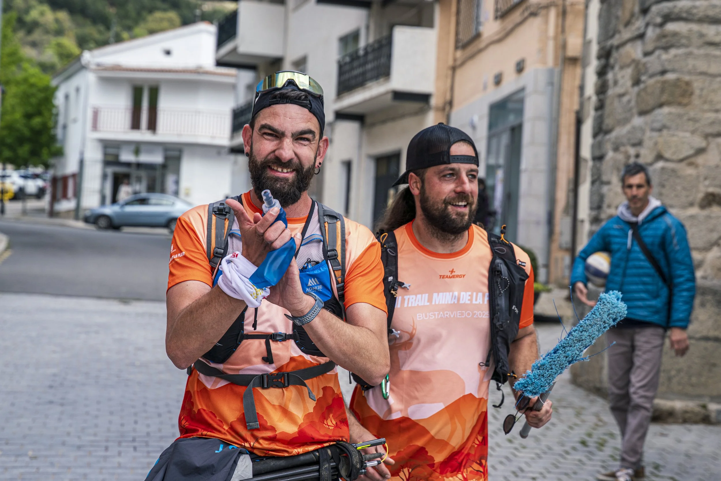 Dos corredores con camisetas naranjasparticipan en una carrera de montaña en una calle urbana. Uno de ellos sostiene un spray para marcar el camino y el otro una escobilla de goma. En el fondo, una persona con chaqueta azul y pantalones claros observ