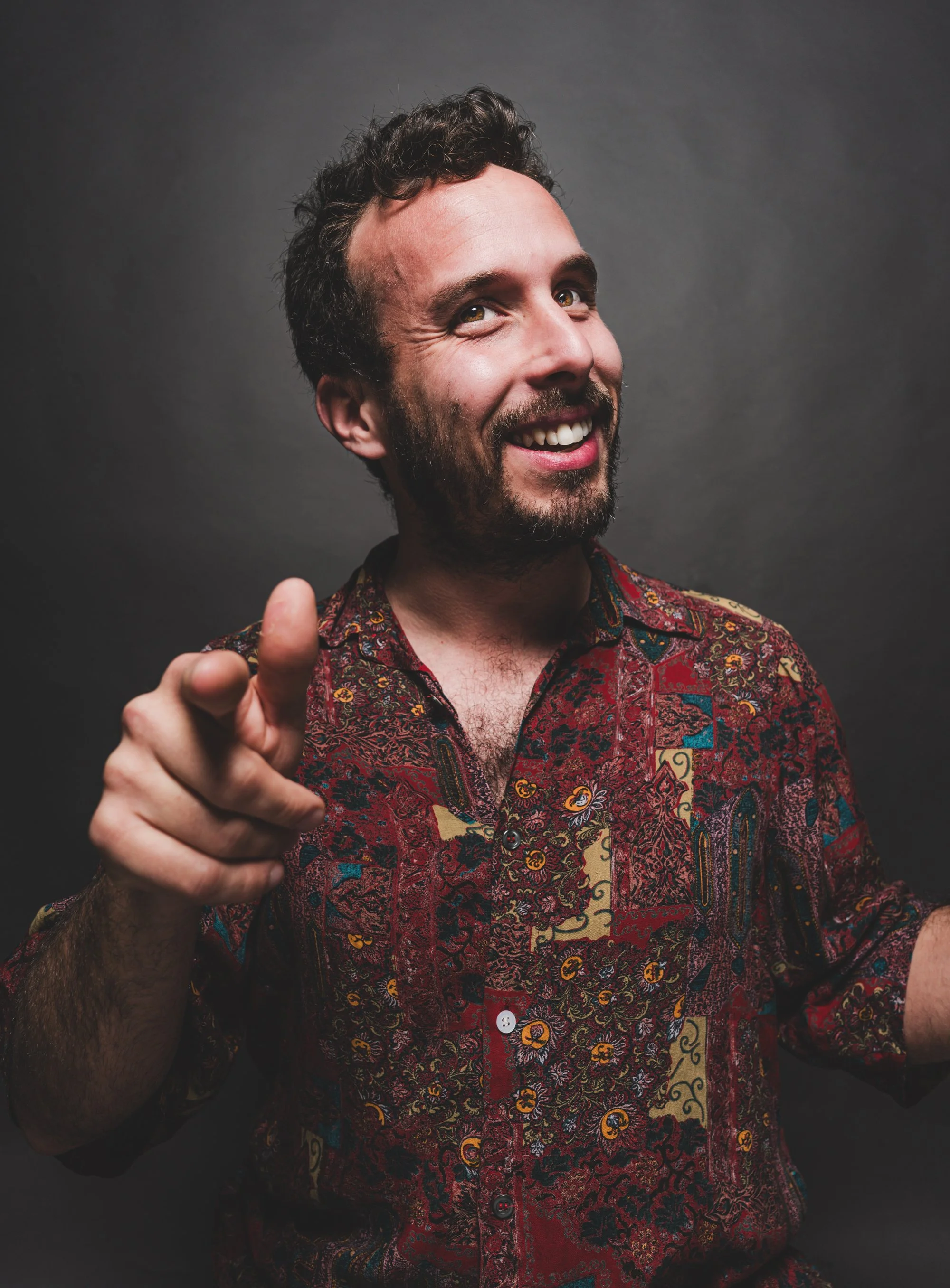 Hombre sonriendo con camisa de colores en un fondo oscuro, haciendo una señal con la mano.
