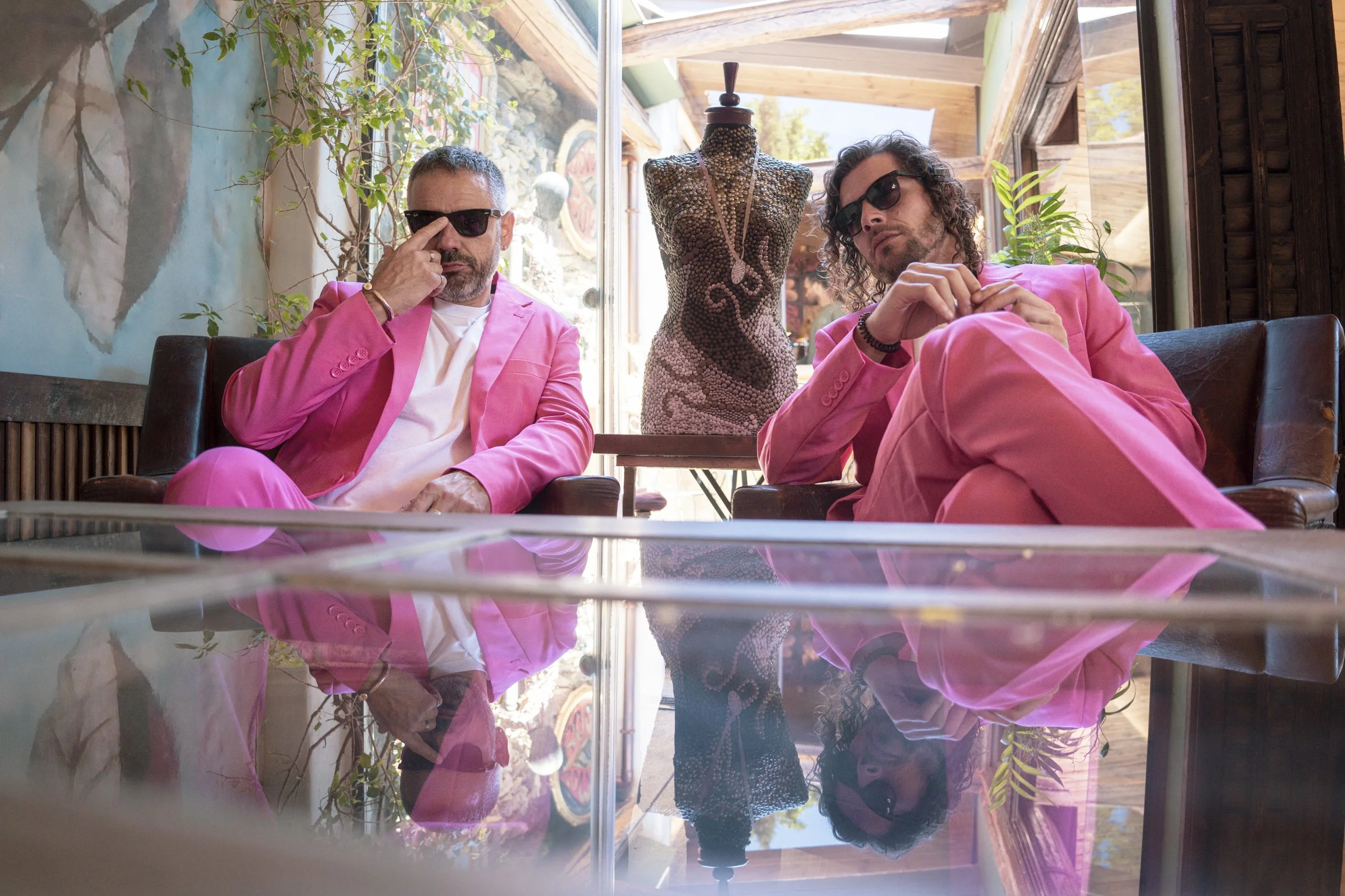 Dos hombres con trajes rosas y gafas oscuras sentados en un sillón en un lugar decorado con plantas y arte, frente a una mesa de cristal que refleja sus caras. Detrás, una estatua de cuerpo entero cubierta de piedras y detalles en el fondo y ventanas