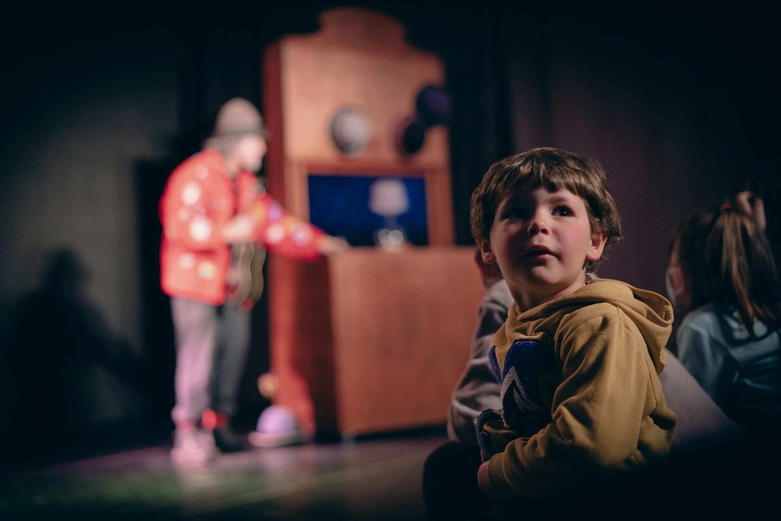 Niño con expresión pensativa observando un espectáculo de teatro con fondo oscuro y un actor en escena.