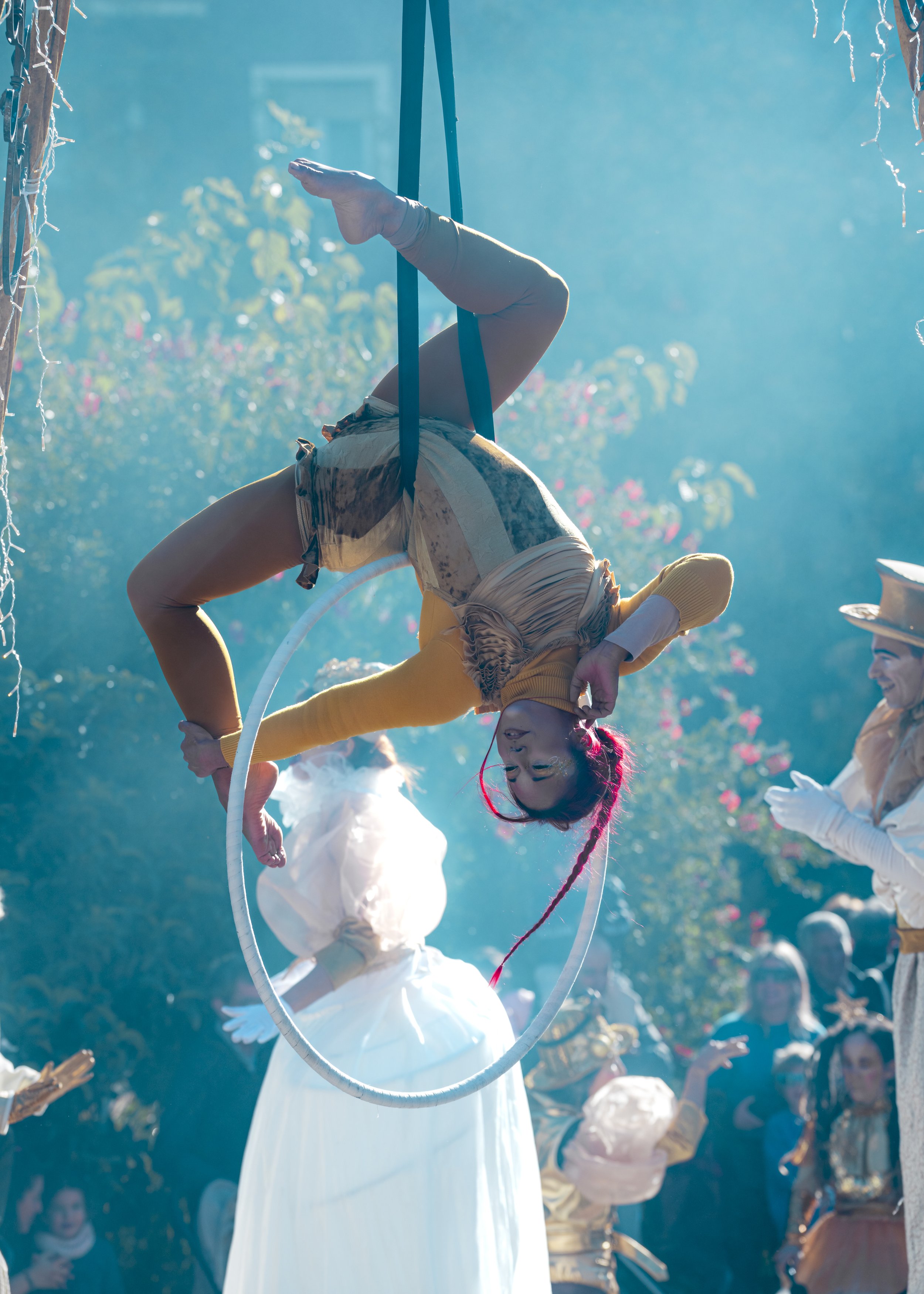 Persona acrobática columpiándose en un aro durante un espectáculo con otras personas disfrazadas de época en el fondo y entre las plantas.