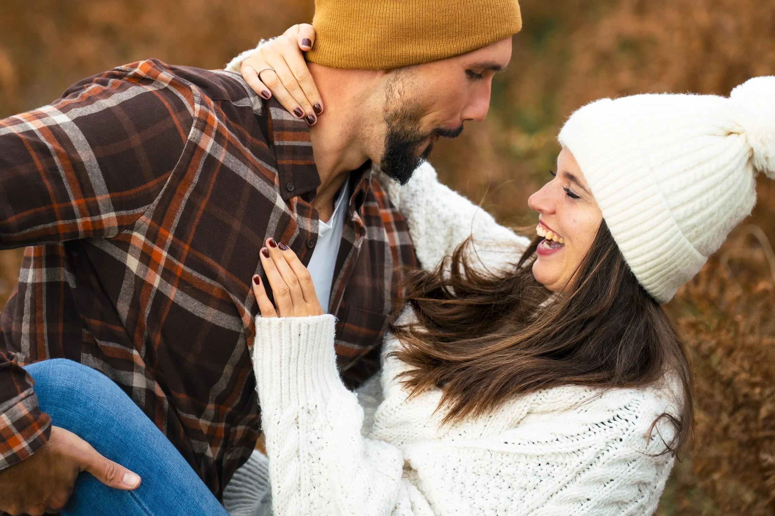 Una pareja divertida y amorosa en un fondo otoñal, la mujer usa suéter blanco y gorro de lana, el hombre lleva una camisa de cuadros y un gorro marrón, ambos están sonriendo y compartiendo un momento romántico y alegre al aire libre.