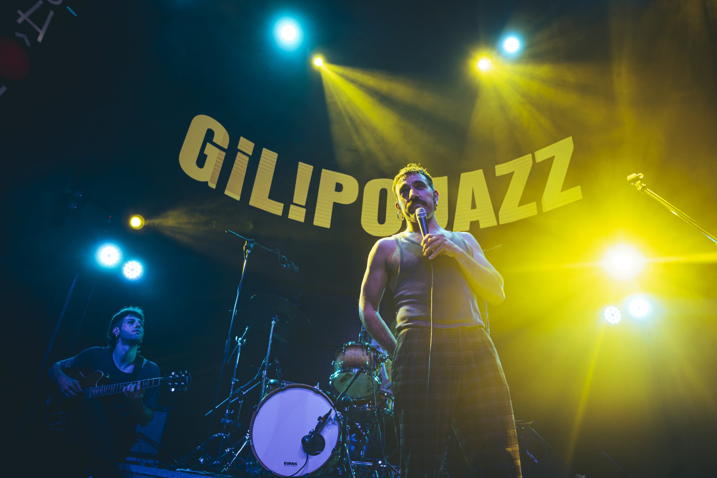 Un cantante con micrófono en mano en un escenario, luces de colores en el fondo, y un músico tocando la guitarra en el lado izquierdo. La palabra 'GILIPOAZZ' aparece en el fondo en letras grandes y amarillas.