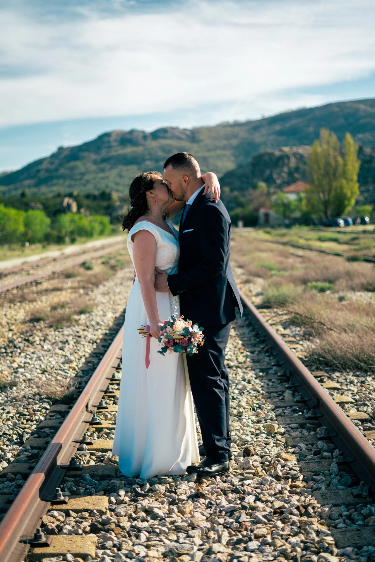 Pareja de novios en una historia de bodas, besándose en las vías del tren en un paisaje de campo y montañas.