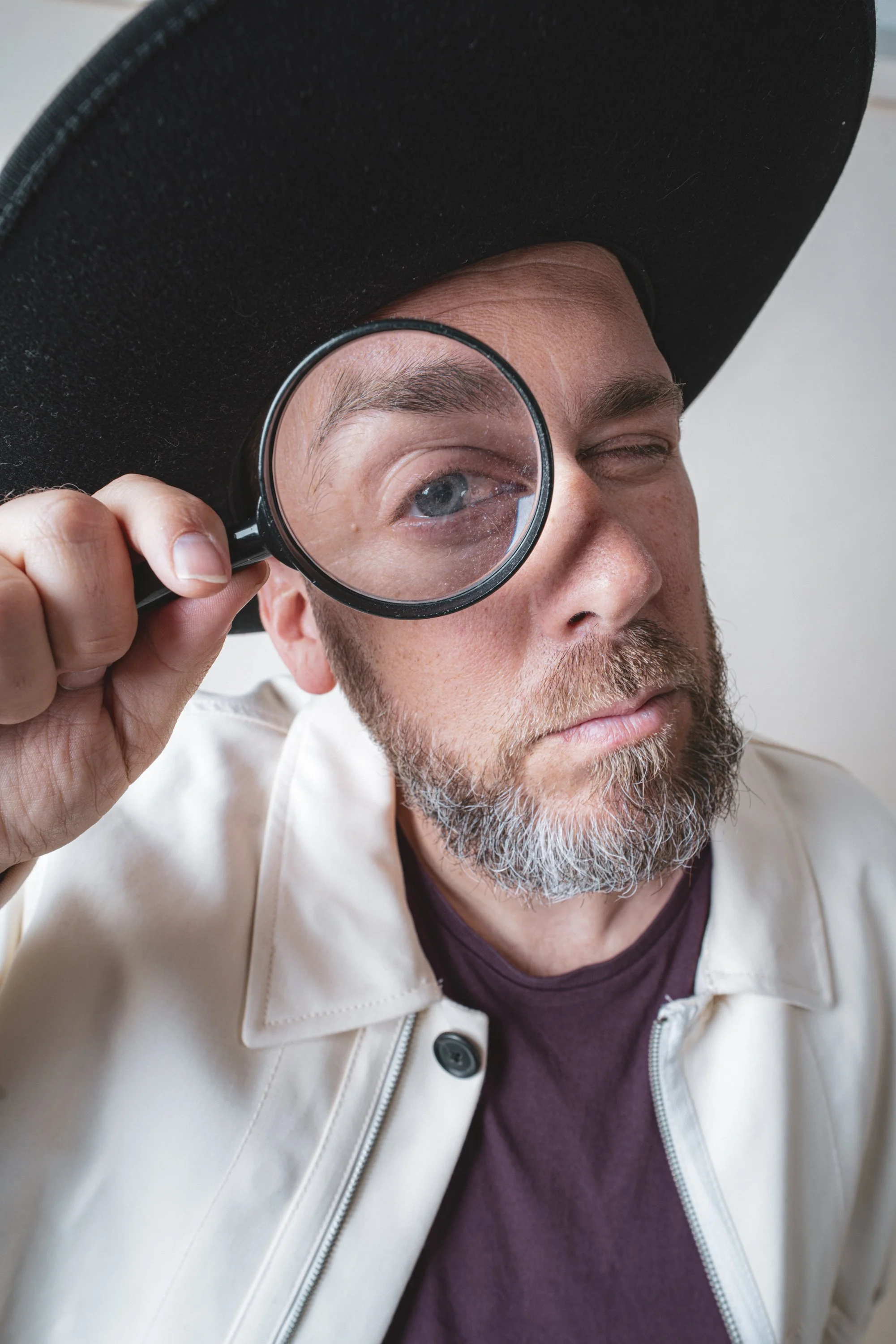 Hombre con barba, usando sombrero grande negro, sostiene una lupa cerca del ojo y parece estar inspeccionando o investigando algo.