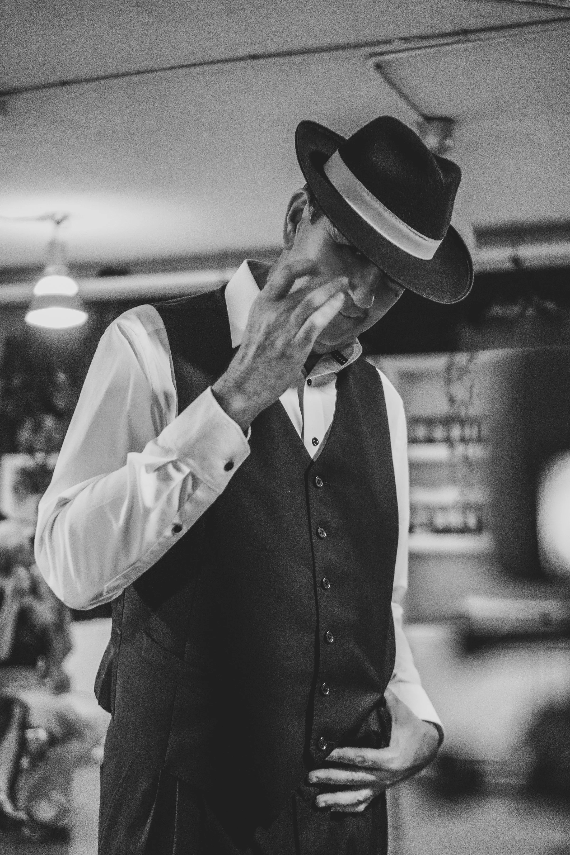 Hombre vestido con camisa blanca, chaleco negro y sombrero oscuro, tocándose la cara con expresión pensativa.