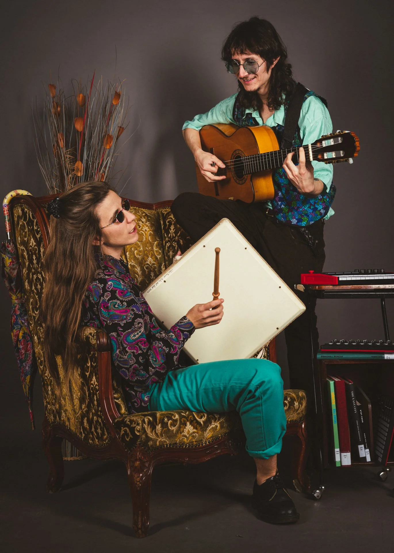Dos músicos jóvenes tocando instrumentos en un estudio. La mujer, sentada en un sillón vintage, sostiene un tambor y lleva gafas de sol. El hombre, de pie sobre el sillón, toca una guitarra y lleva gafas oscuras.